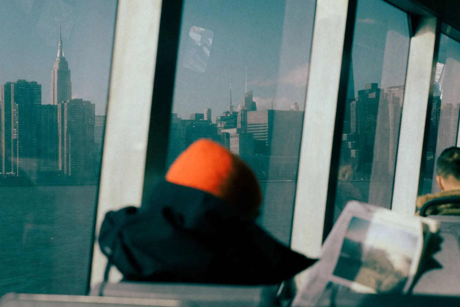 View through an airplane window showing New York City skyline, including the Empire State Building, with passengers seated nearby, one reading a tablet and another wearing an orange hat.