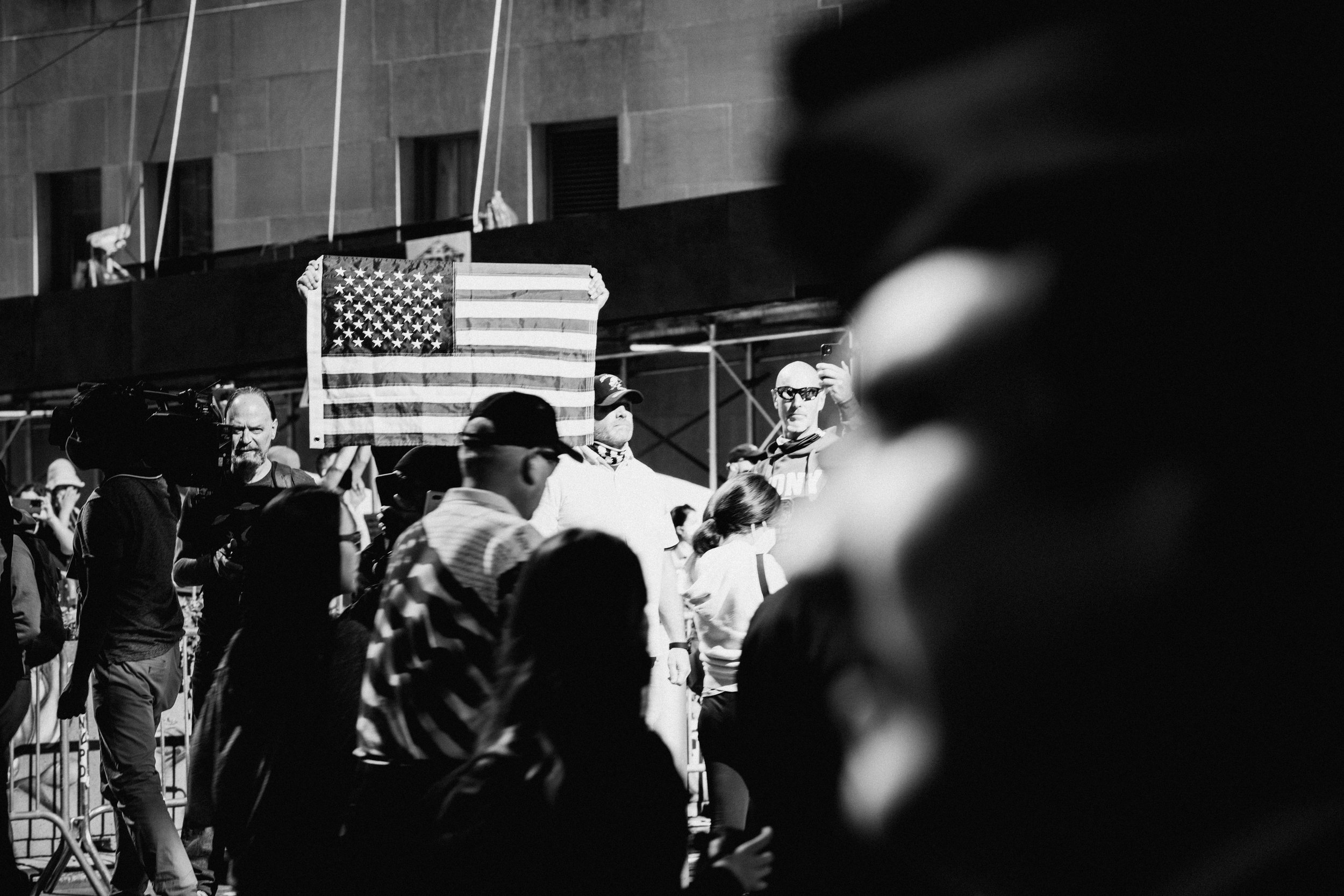 Black and white photo of a crowd at a protest or rally, with some individuals holding signs or flags. A person in the background holds an American flag. The scene appears to be outdoors, with a building in the background.