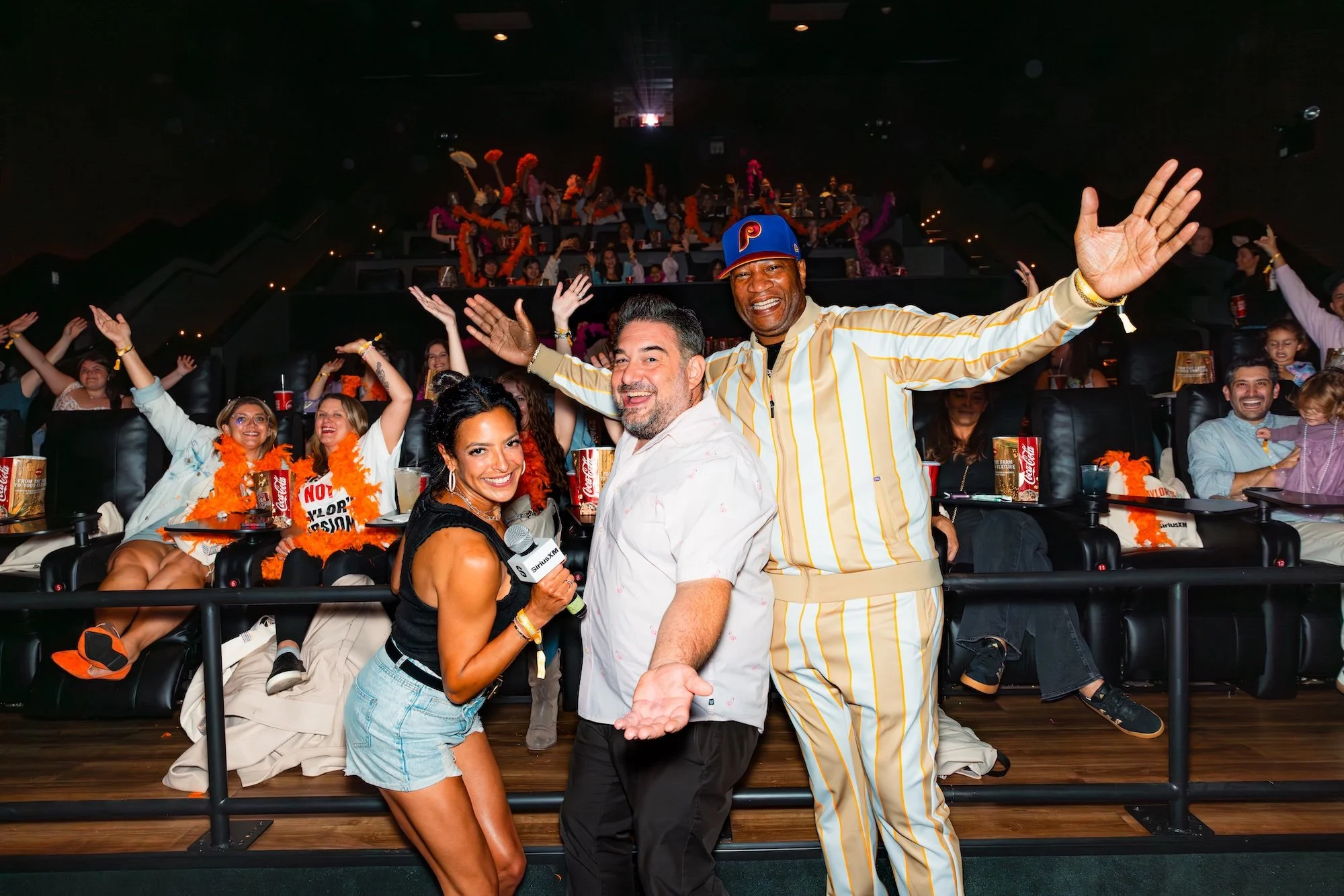 People at an event or show, with three individuals in the front smiling and posing for the camera. The woman in the front is holding a microphone and wears a black top and denim shorts, while the man in the middle has a beard and is wearing a white s