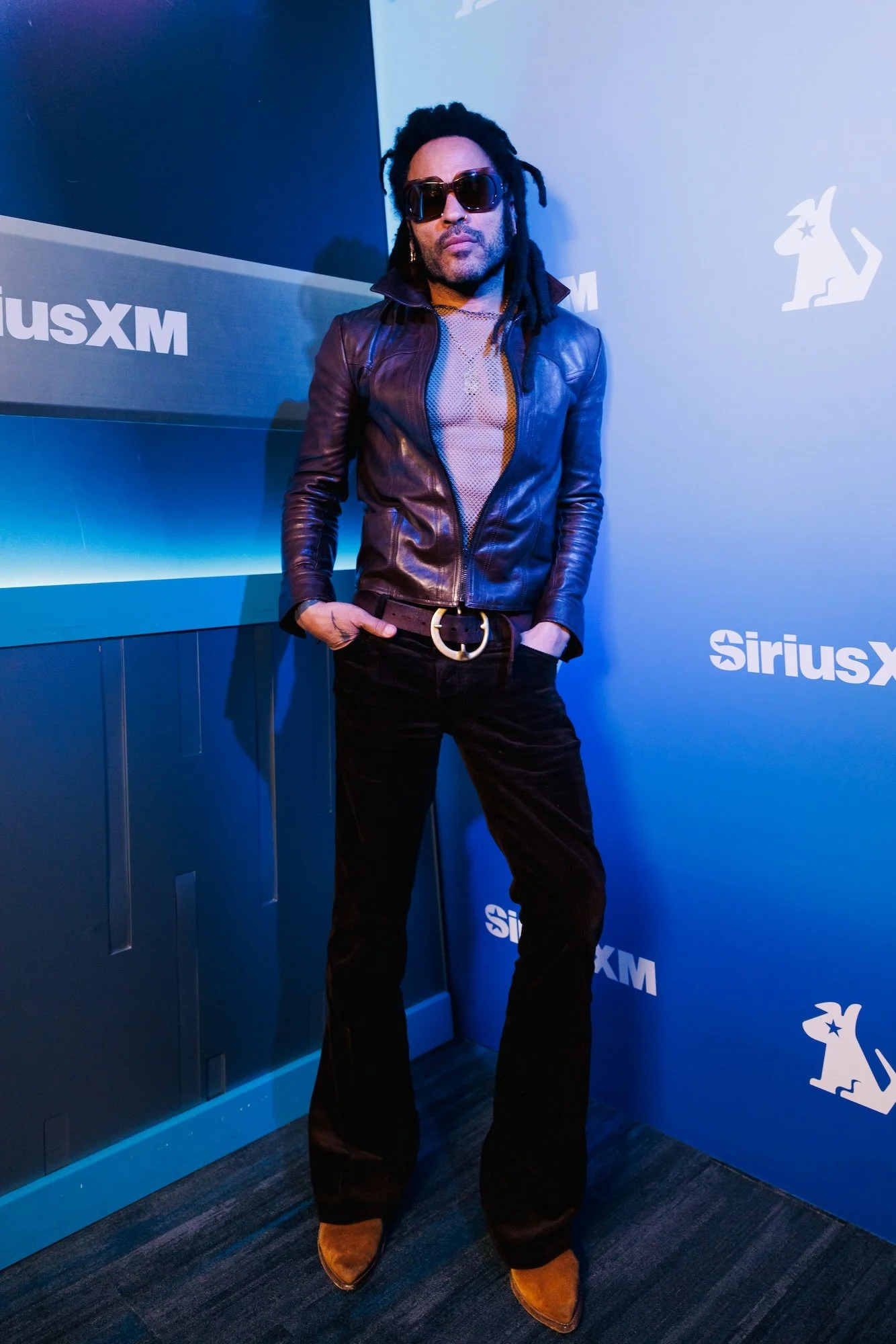 Man with dreadlocks wearing sunglasses, a leather jacket, black pants, and brown boots, standing in front of a SiriusXM and Kangaroo logo background.