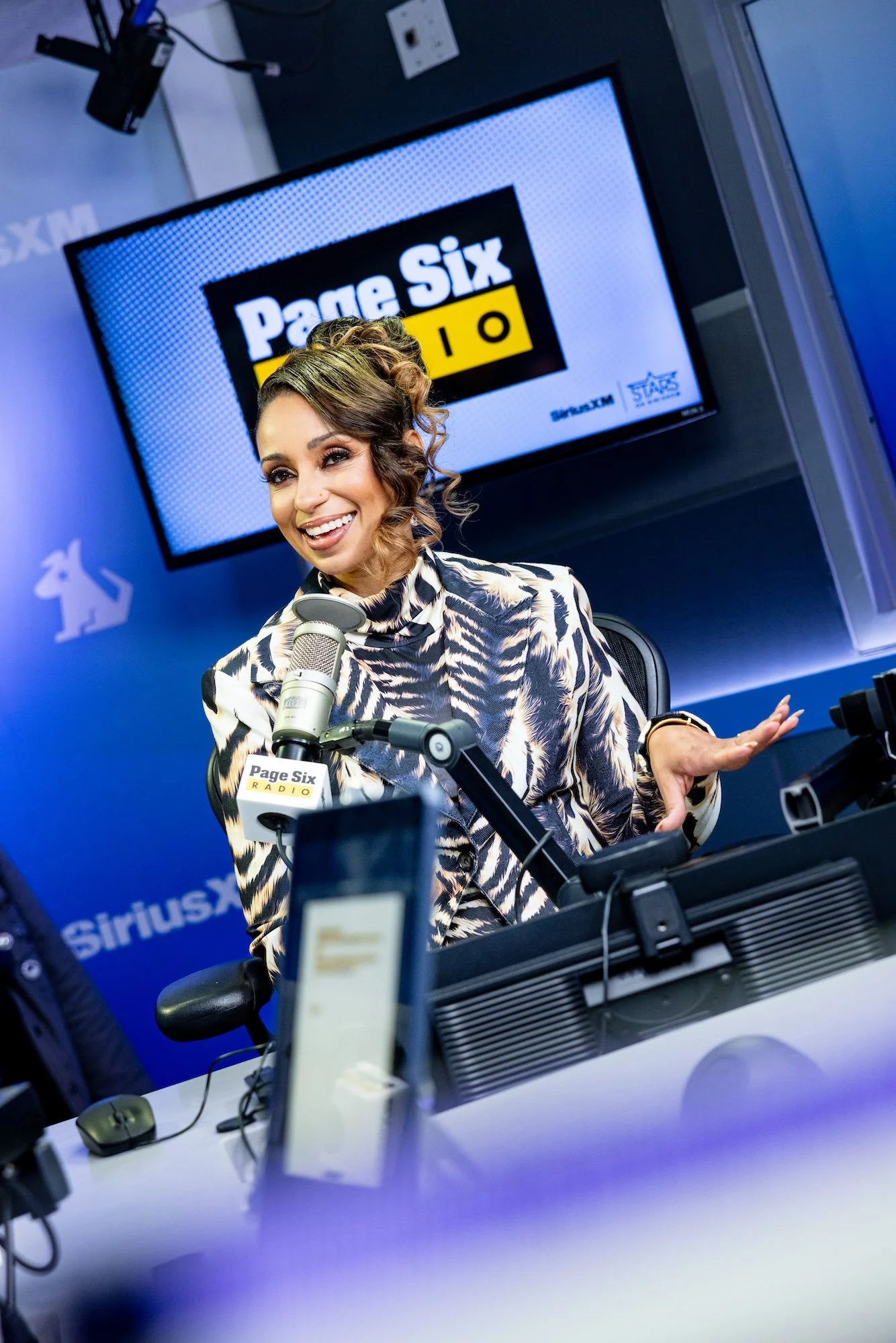 A woman with styled hair and makeup smiling and speaking into a microphone during a radio show at Page Six Radio studio, with a screen in the background displaying the Page Six Radio logo.
