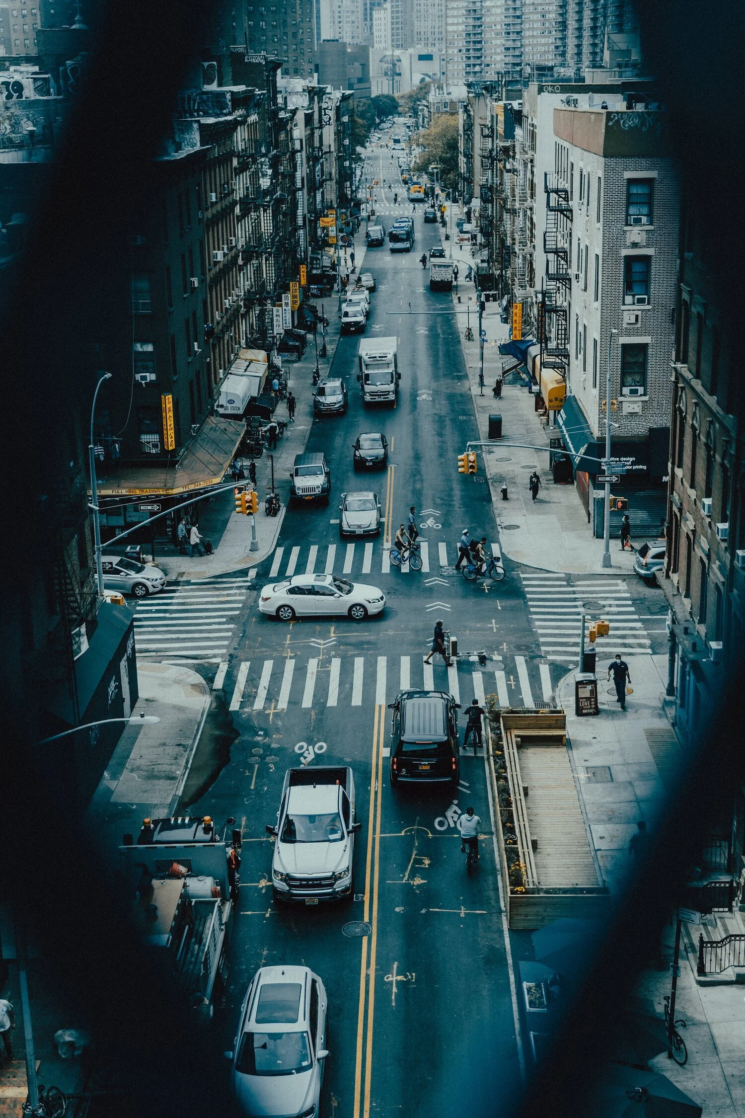 City street view seen through window with some black framing. Traffic includes cars and delivery trucks, with crosswalks and bike lanes. High-rise buildings line the street.
