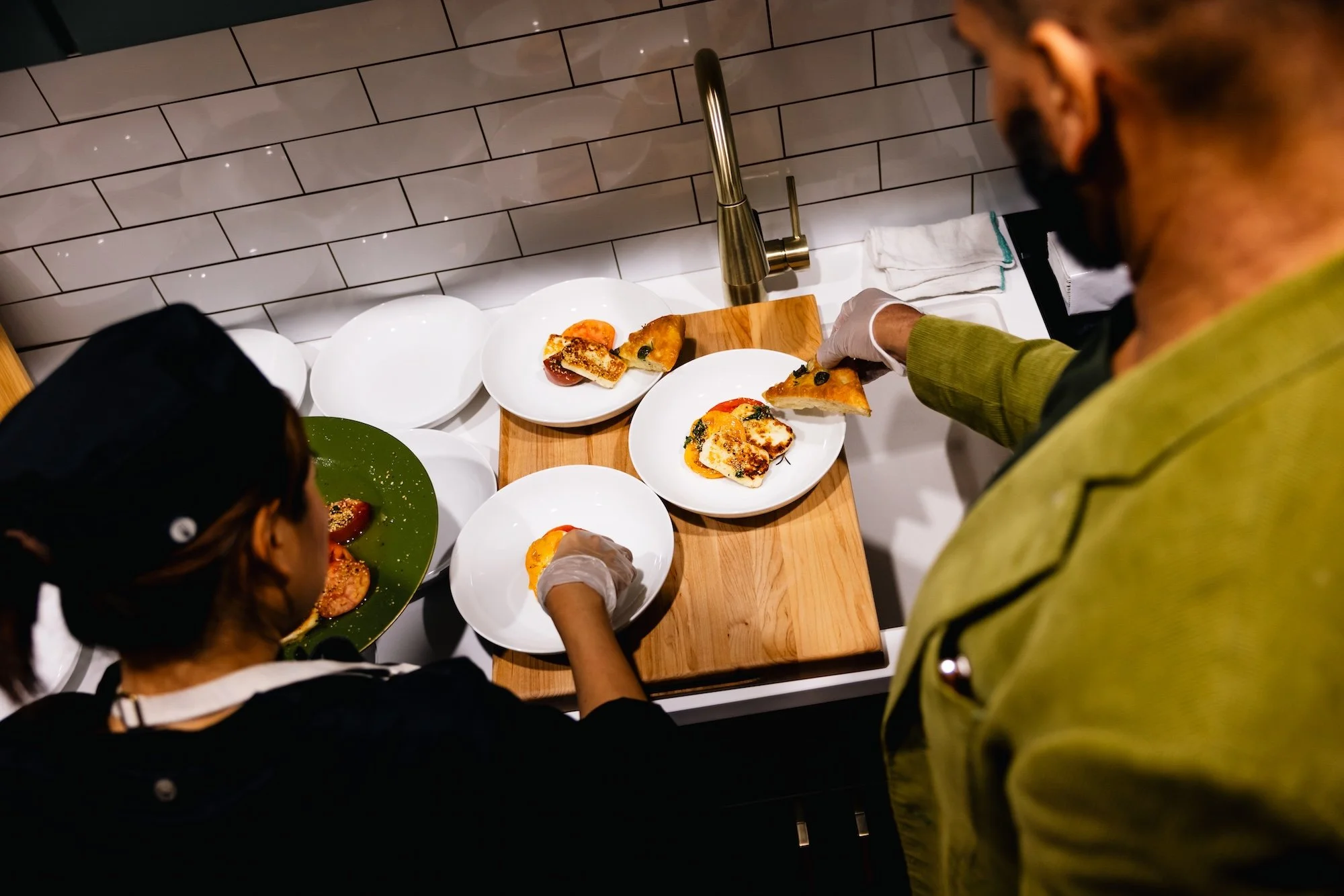 Two chefs preparing and plating slices of pizza in a modern kitchen with white subway tile backsplash.