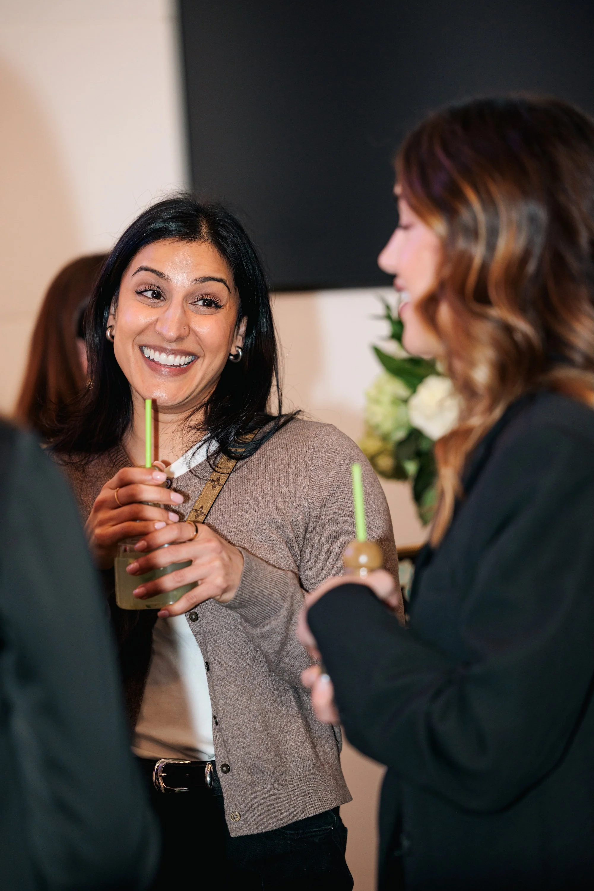 Two women with drinks, smiling and talking in a social setting, with a blurred background of flowers.