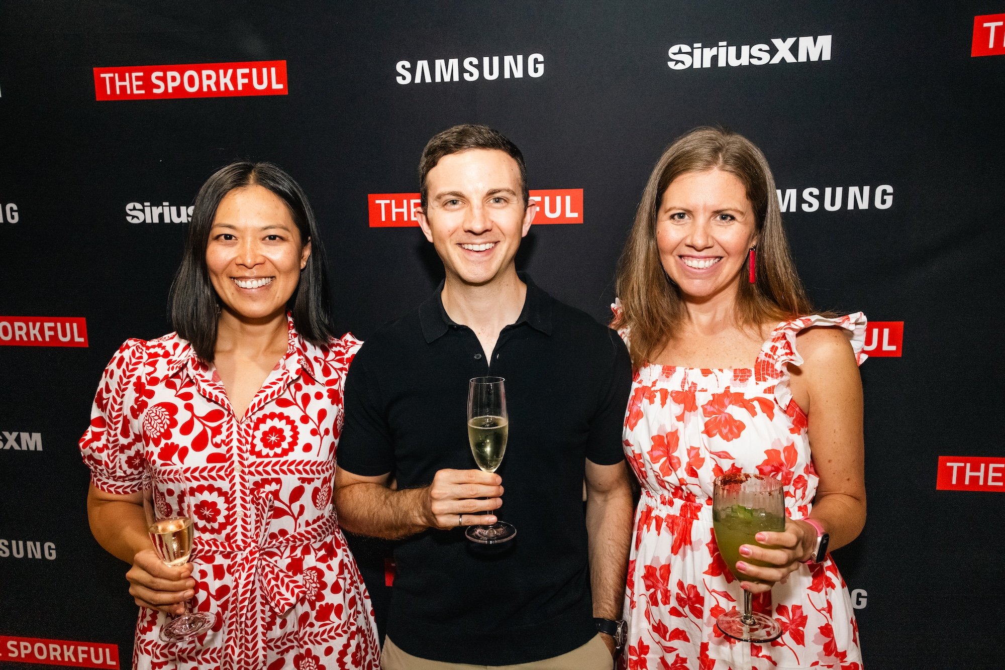 Three people smiling at a social event, holding drinks in front of a black backdrop with logos for 'The Sporkful', 'SiriusXM', and 'Samsung', with two women on either side of a man in a black polo shirt.