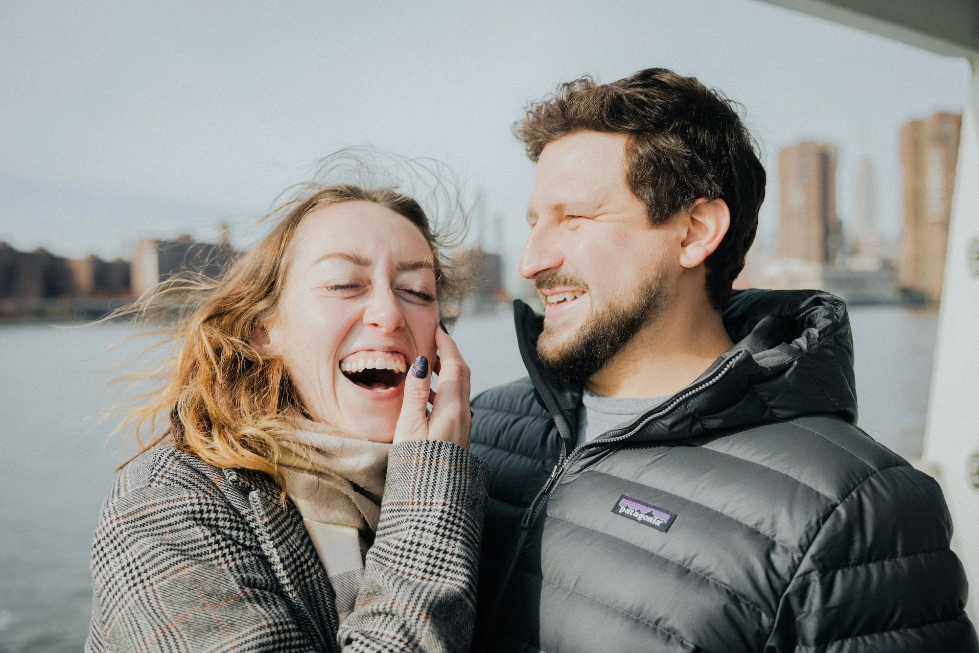 A woman and a man are smiling and laughing near a body of water with city buildings in the background.