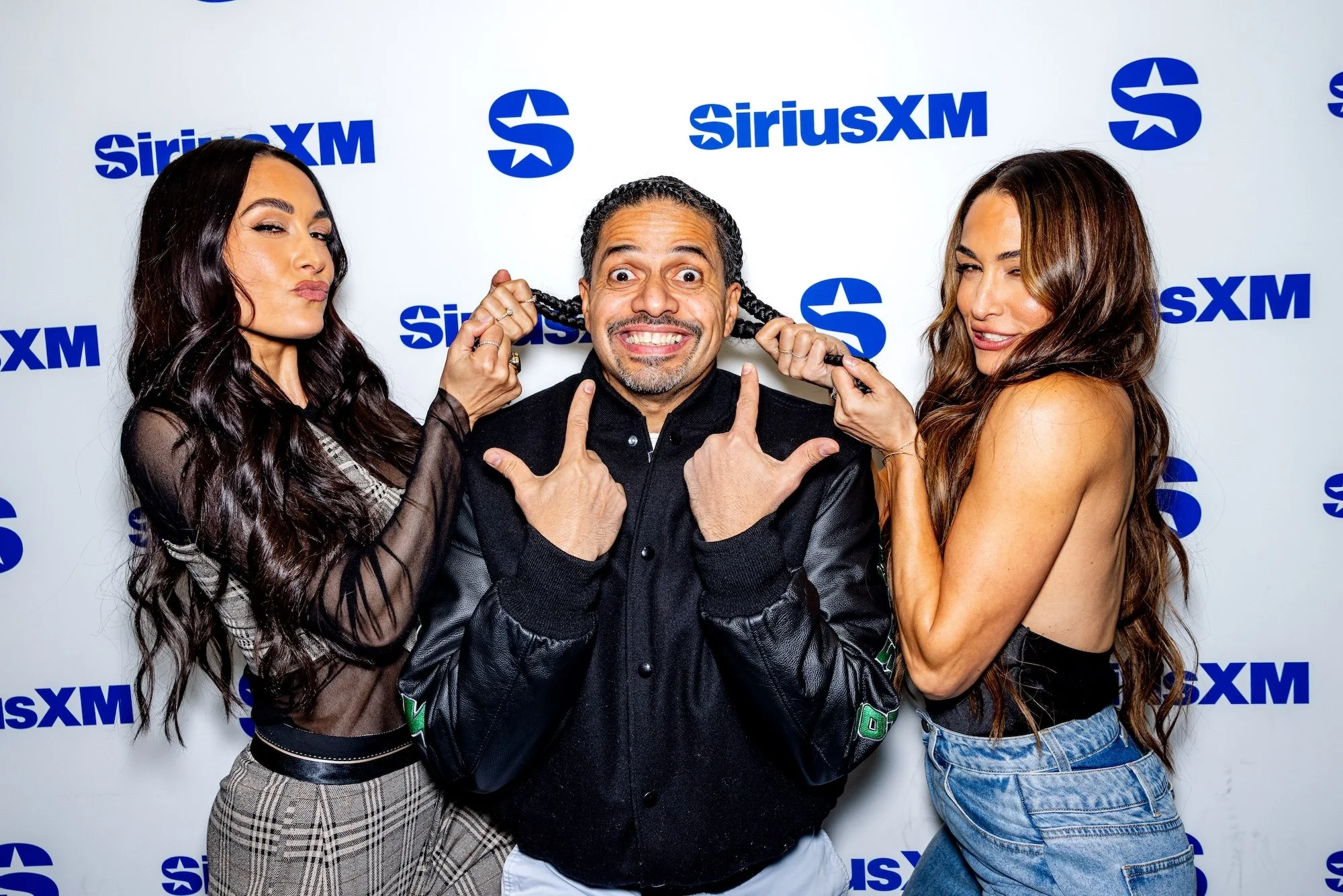 Two women playfully tug at a man's braided hair in front of a SiriusXM backdrop. The man is smiling widely and making a rock-and-roll hand gesture.