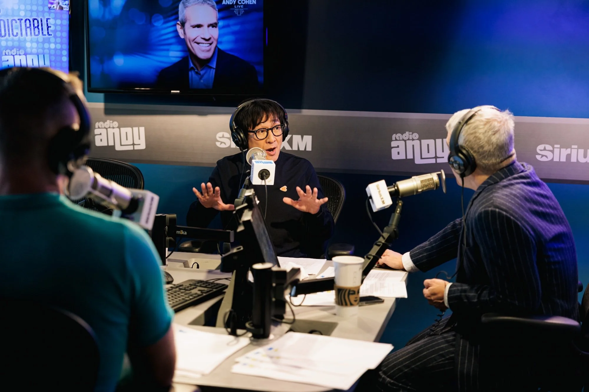 People conducting a radio interview in a studio with microphones, headphones, papers, and a computer on the table. A large screen behind them shows a smiling man.