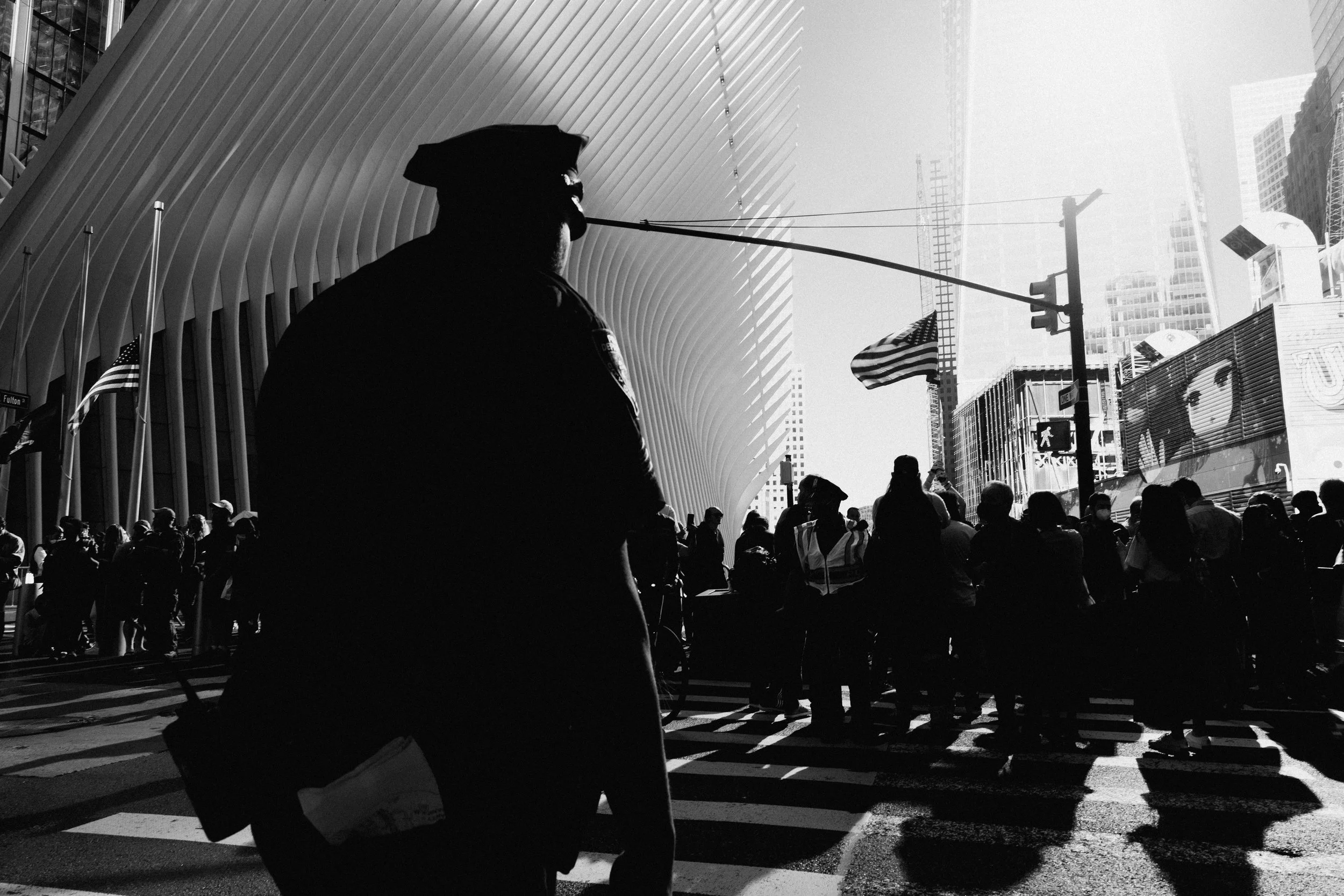 A black and white photo of a police officer standing at a busy crosswalk in an urban area, with a crowd of people crossing the street and city buildings in the background.
