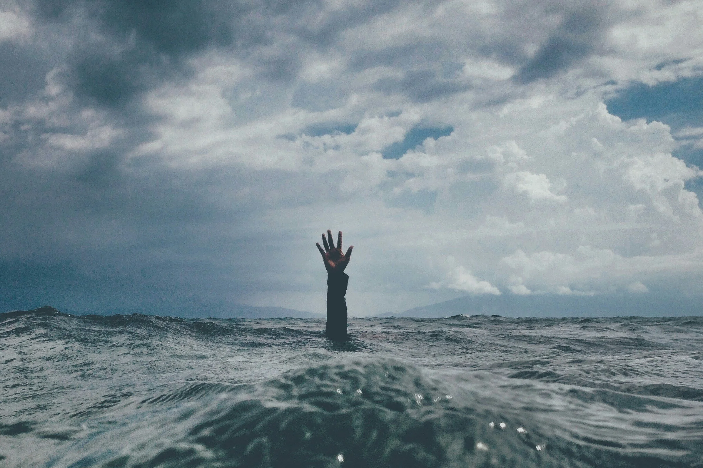 A person is in the water with only their arm and hand visible, reaching out towards the sky, under a cloudy sky.