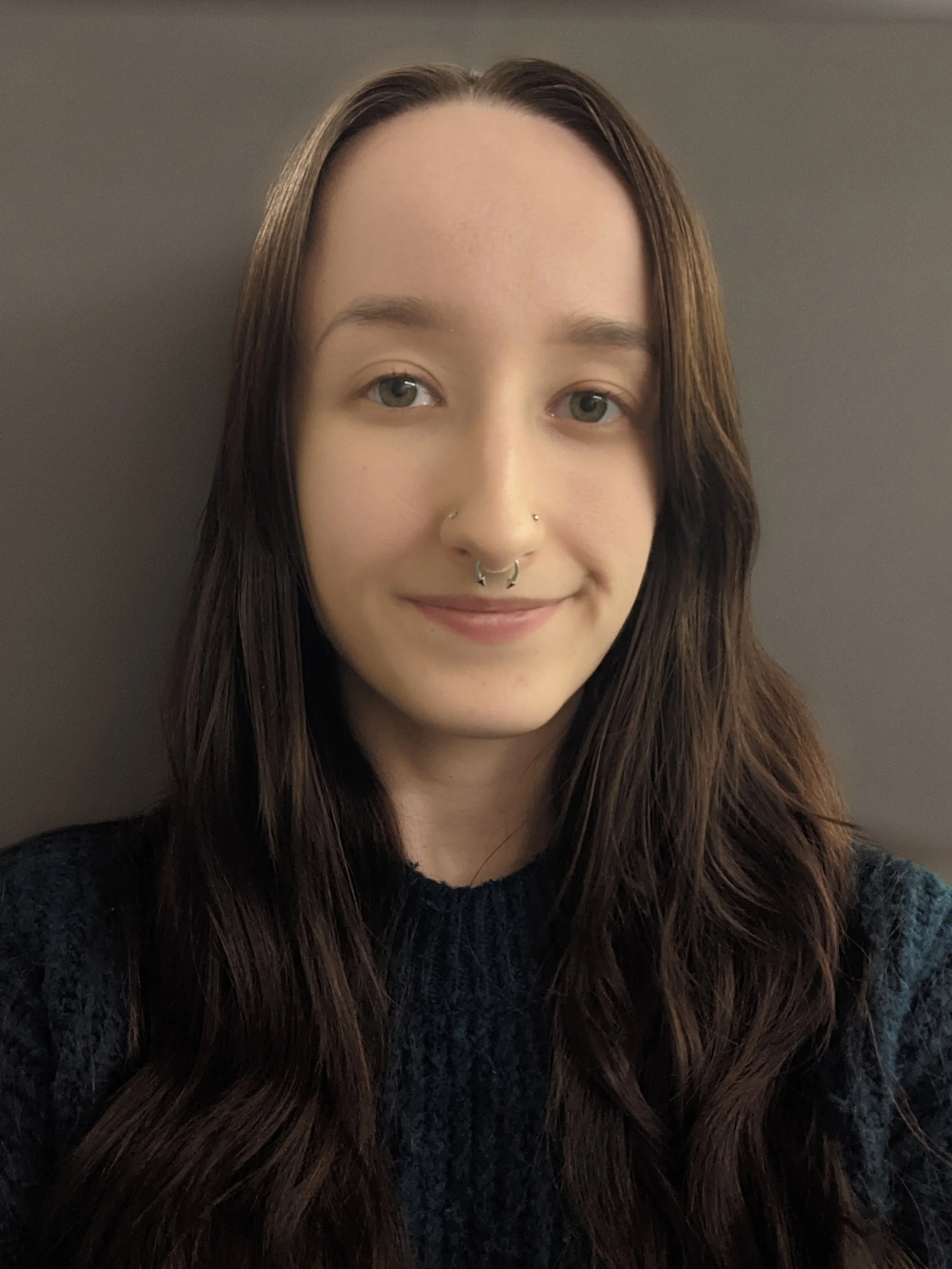 A young woman with long brown hair and light skin takes a selfie against a plain, neutral-colored wall. She has a septum piercing and is smiling softly.