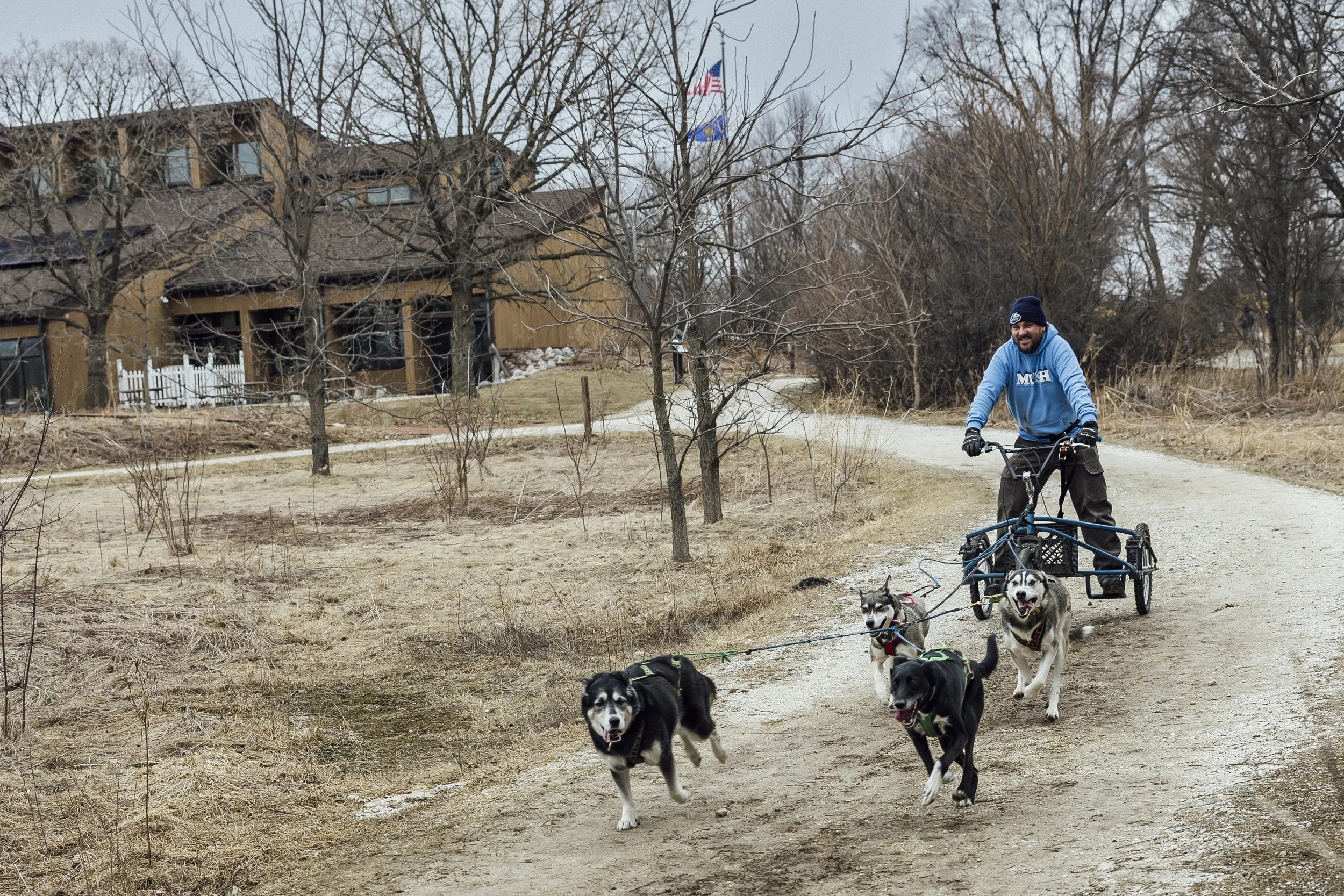A team of sled dogs running through the snow, the sled with a female musher driving it, and another person riding in the basket.