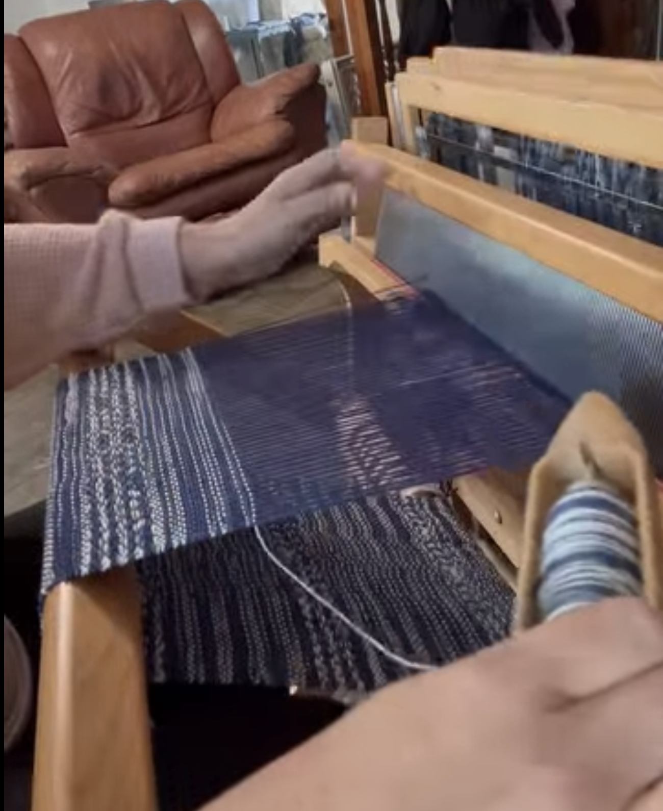 Image of a weaving loom, and a pair of hands. One hand has the shuttle with the thread in it, and the other hand is operating the loom. On the loom is an indigo and white woven project.
