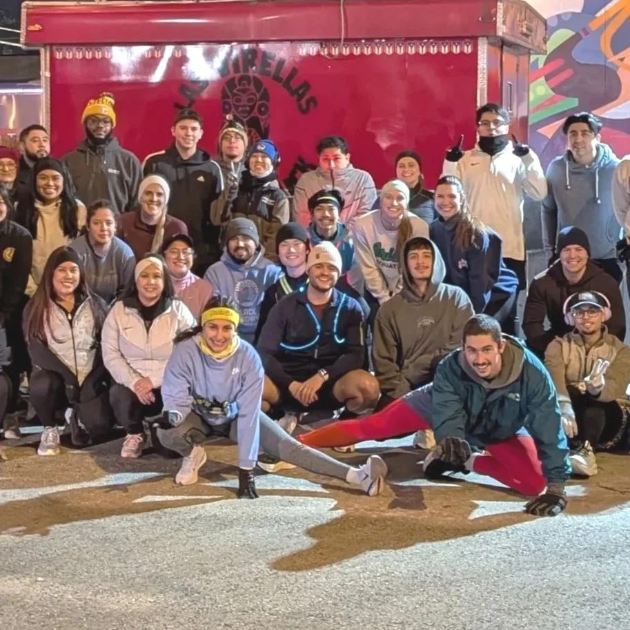A huge group of outdoor runners are posed in a group photo, with a few of them sporting headbands and hats, and a few in the front doing runners stretches.