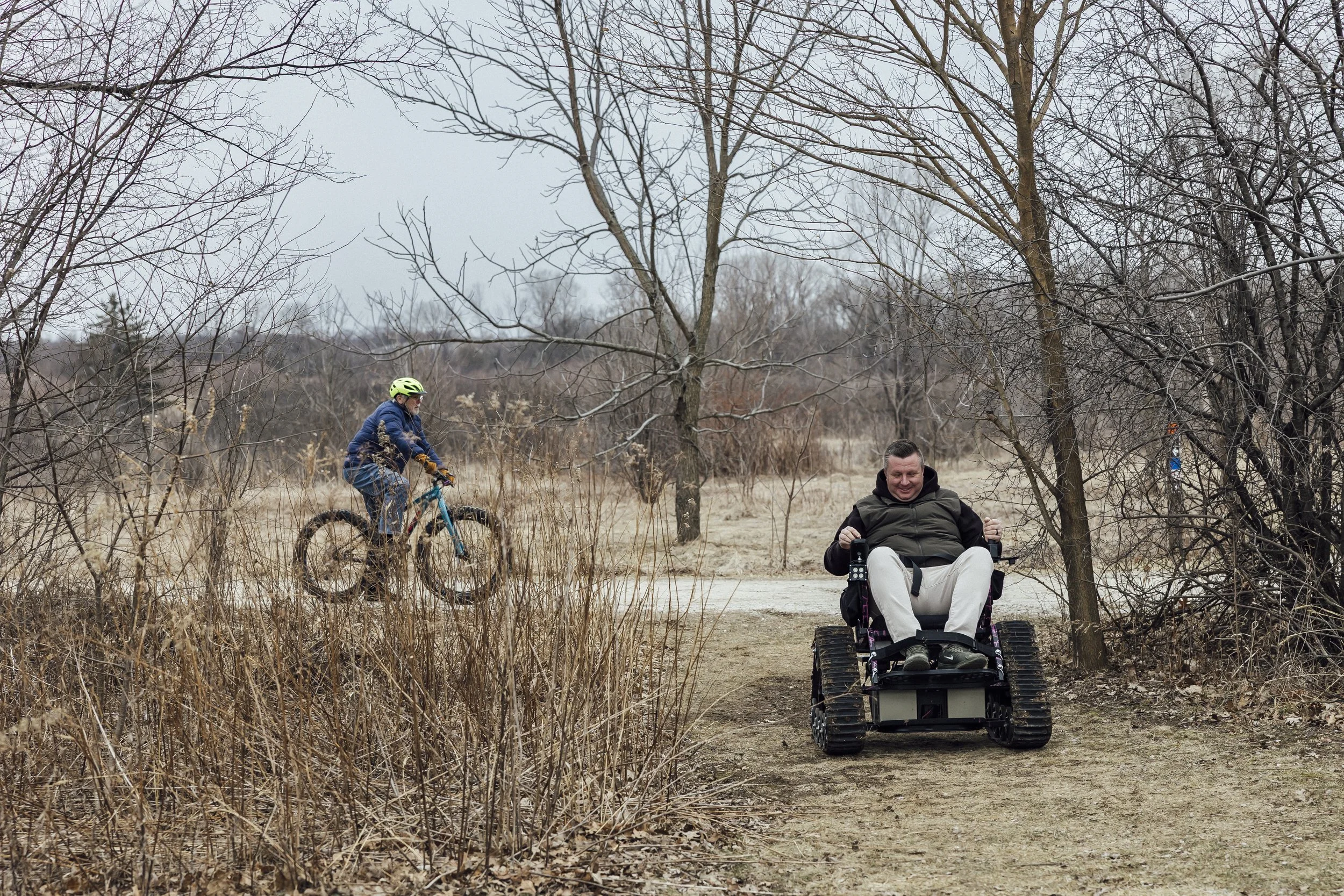 An outdoor scene with a man in the background biking, and a man in the foreground sitting on an all-terrain wheelchair. The wheelchair looks like a powered chair, but instead of wheels, it has treads on a belt like a mini tank. 