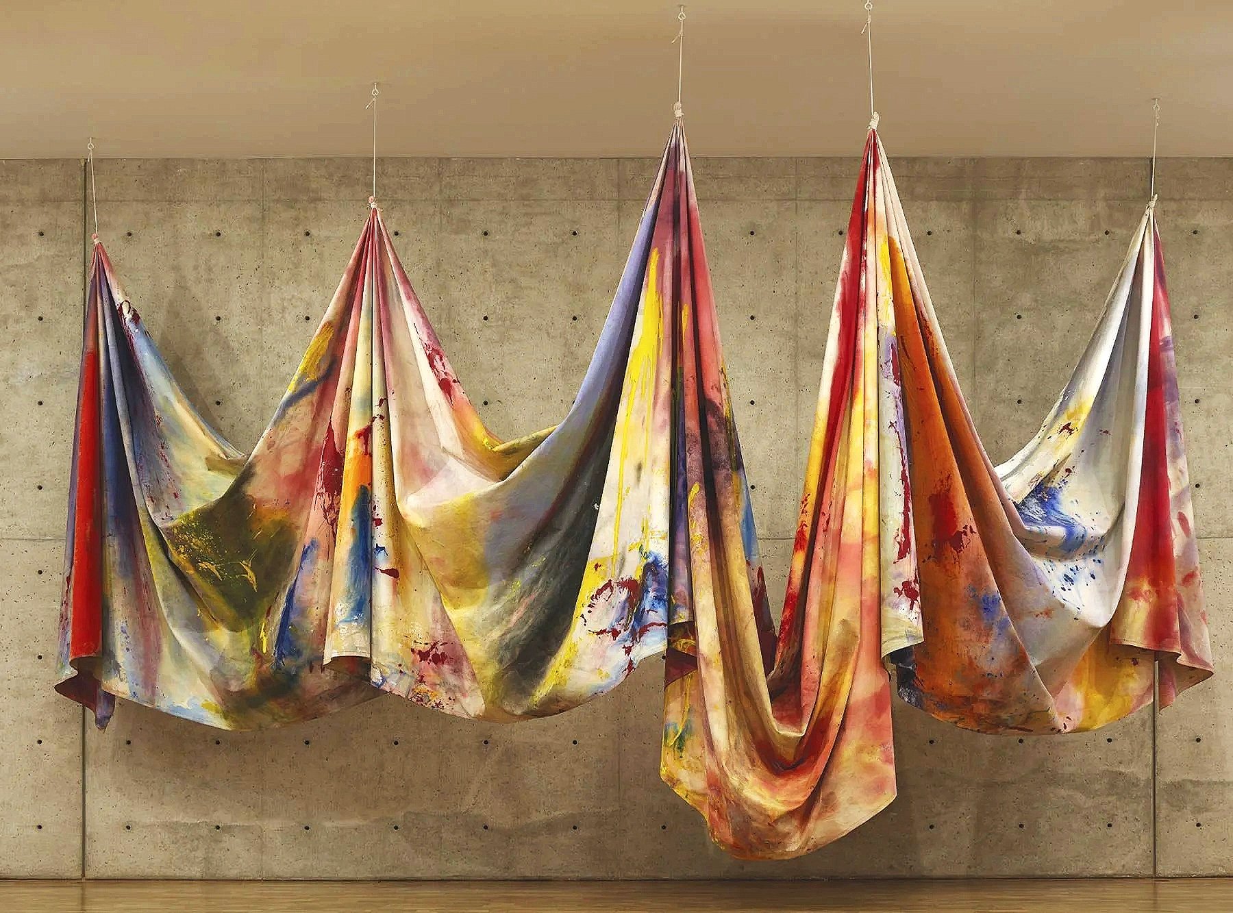 A piece of art by Sam Gilliam called Carousel Merge 2. It is painted canvas draped, and hung from the ceiling in gathers. The paint on the canvas is multi colored: red, blue, yellow mostly. 