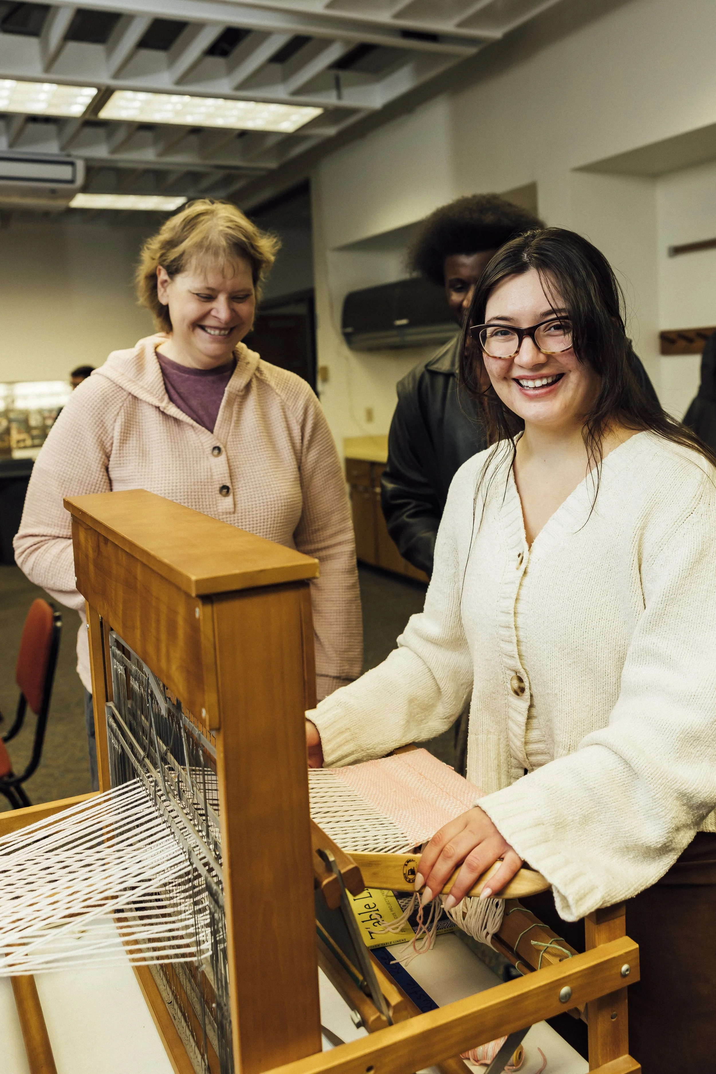 Image of a weaving loom, and a pair of hands. One hand has the shuttle with the thread in it, and the other hand is operating the loom. On the loom is an indigo and white woven project. 