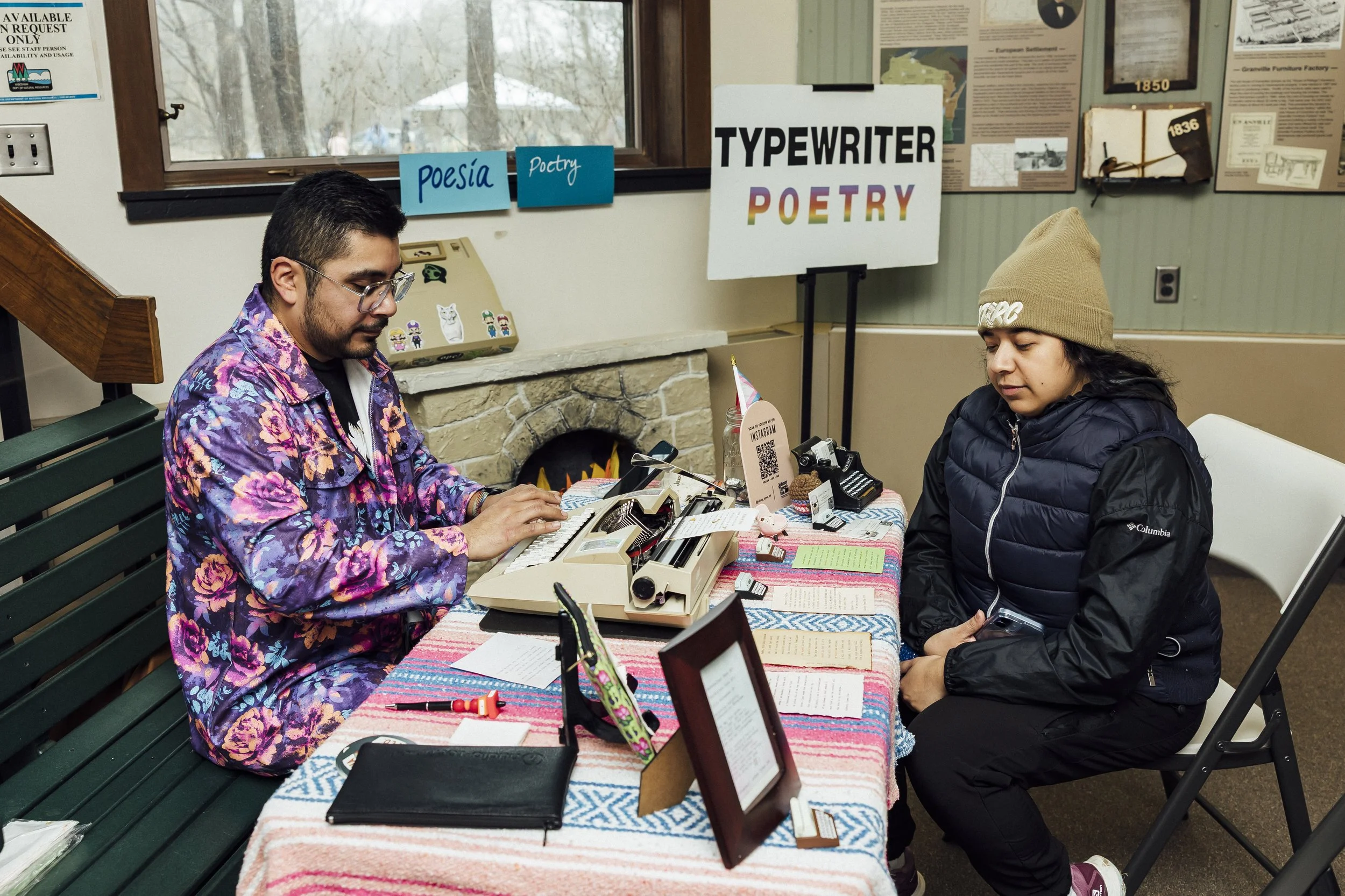 A smiling man in a cap and scarf sits at a table with a typewriter. The sign behind him says 'typewriter poetry'.