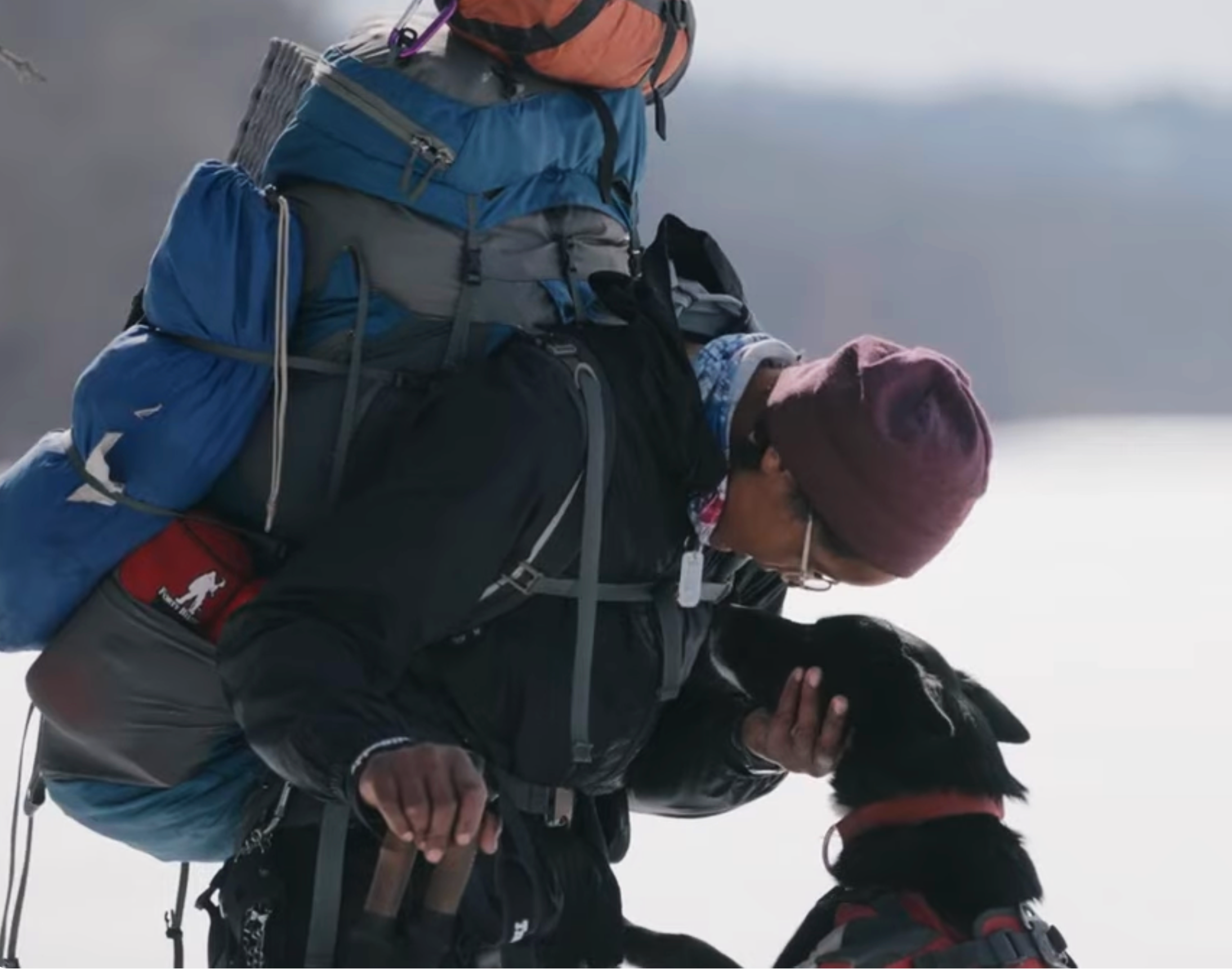 A woman pulling her hood over her ears, standing in the snow with her dog. The black dog is standing on its hind legs and has its front paws on her abdomen.