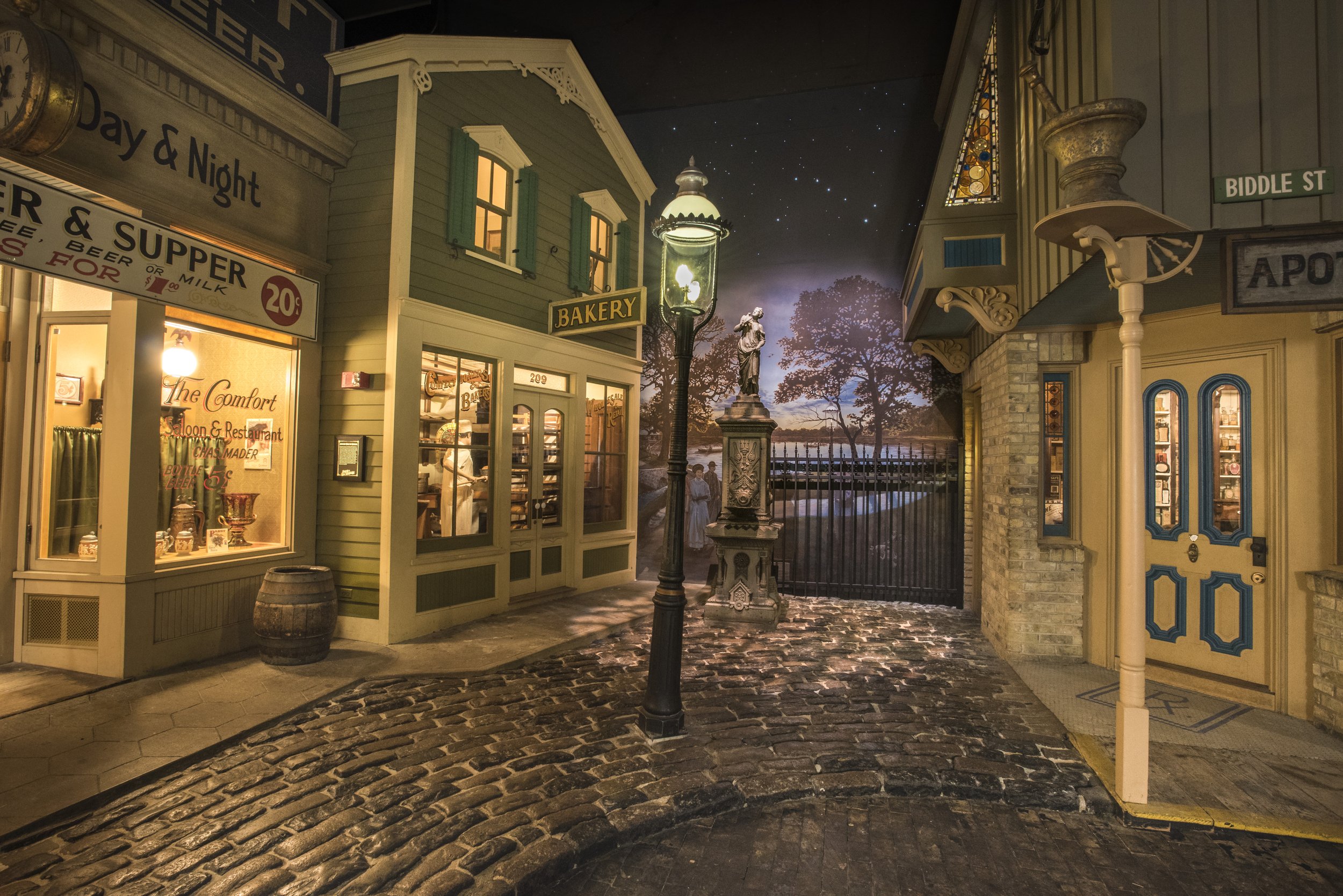 A streetscape inside the Milwaukee Public Museum of the Streets of Old Milwaukee, a recreation of Milwaukee in the early 1900s. You can see a cobblestone street with a bakery, a restaurant, and a fountain. 