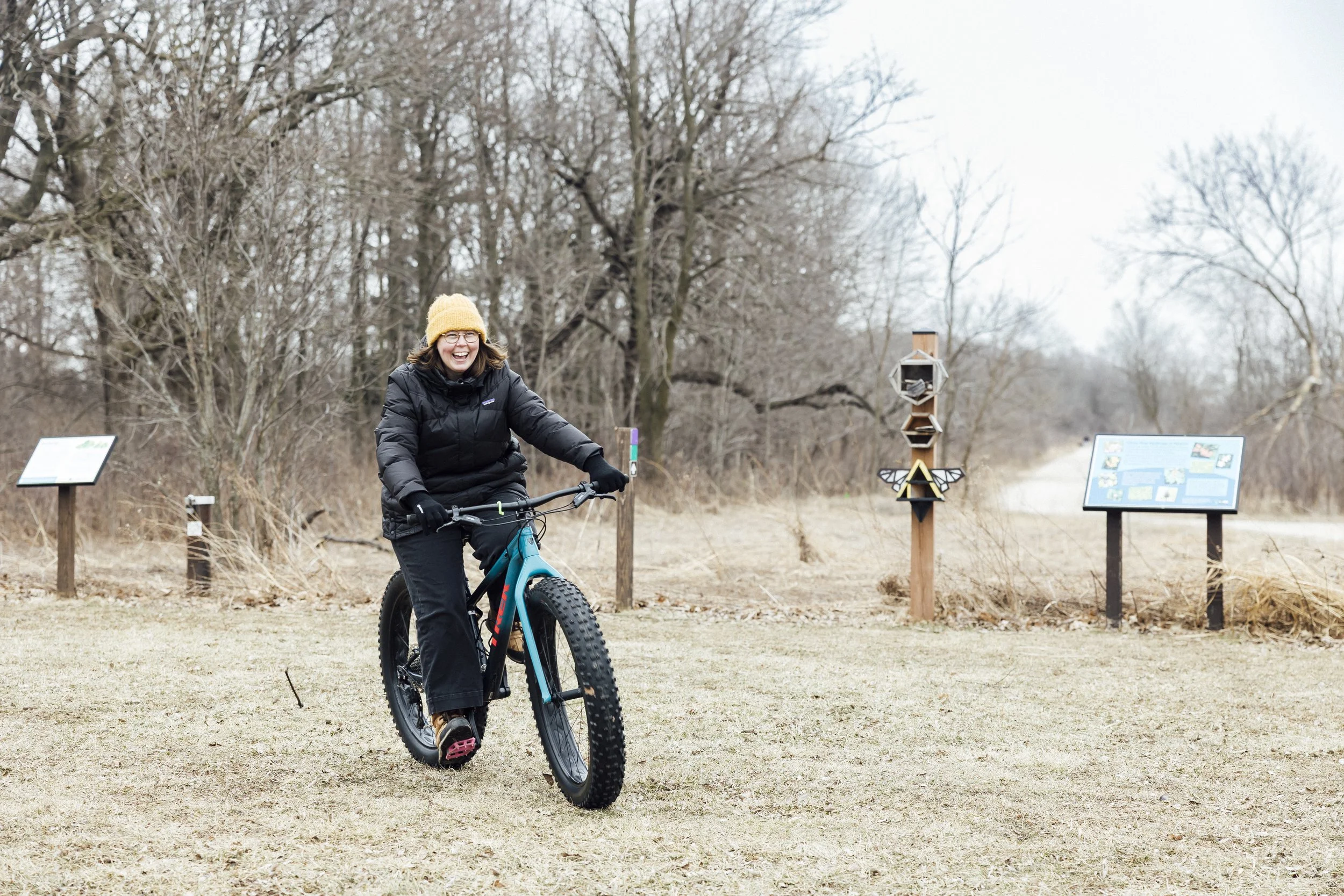 A woman on a fat tire bike pedals over a lawn. She has a yellow hat and a joyous face on. 