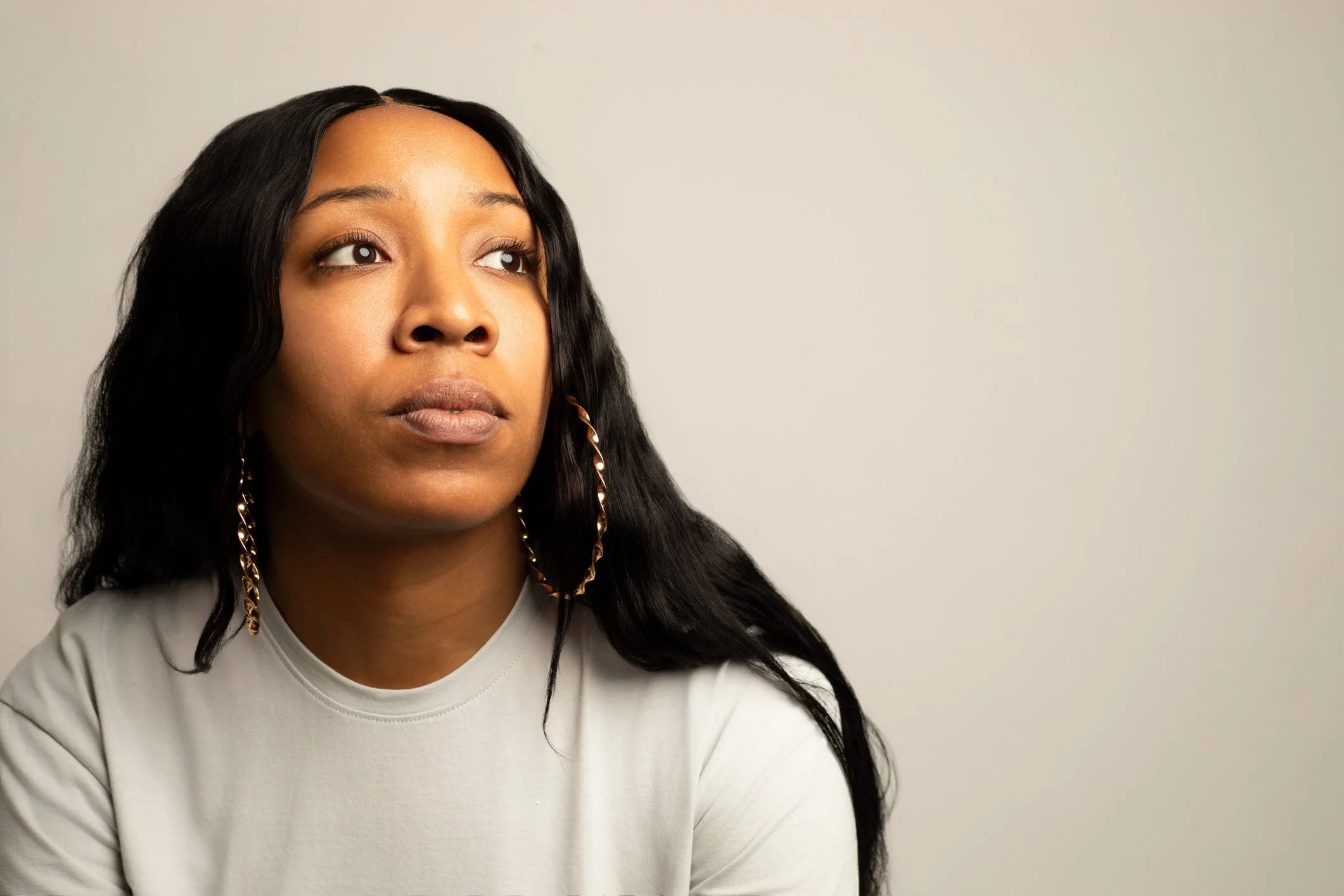 A young woman who was sexually abused with long black hair, wearing large gold hoop earrings and a light-colored top, looking slightly upward against a plain background as she consideres a healing study by Trees of Hope