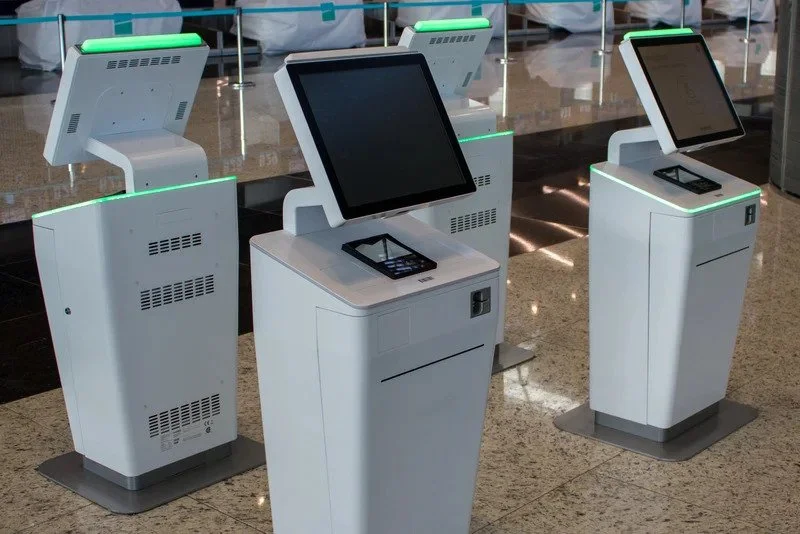 Row of modern self-service check-in kiosks with touchscreen displays and green lights at an airport terminal