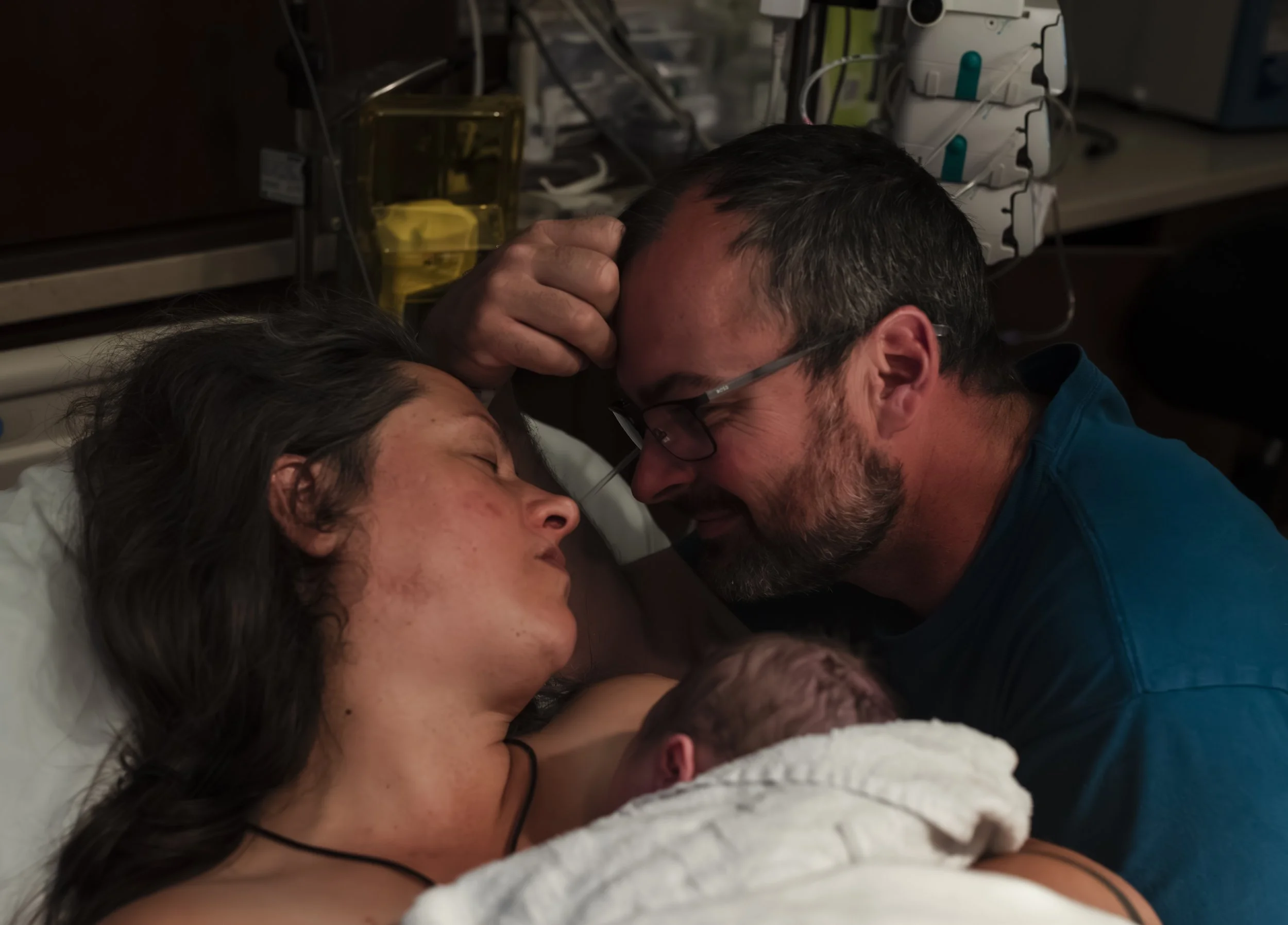 A couple lying in a hospital bed, touching foreheads, with the woman asleep and the man looking at her with concern in a hospital room.