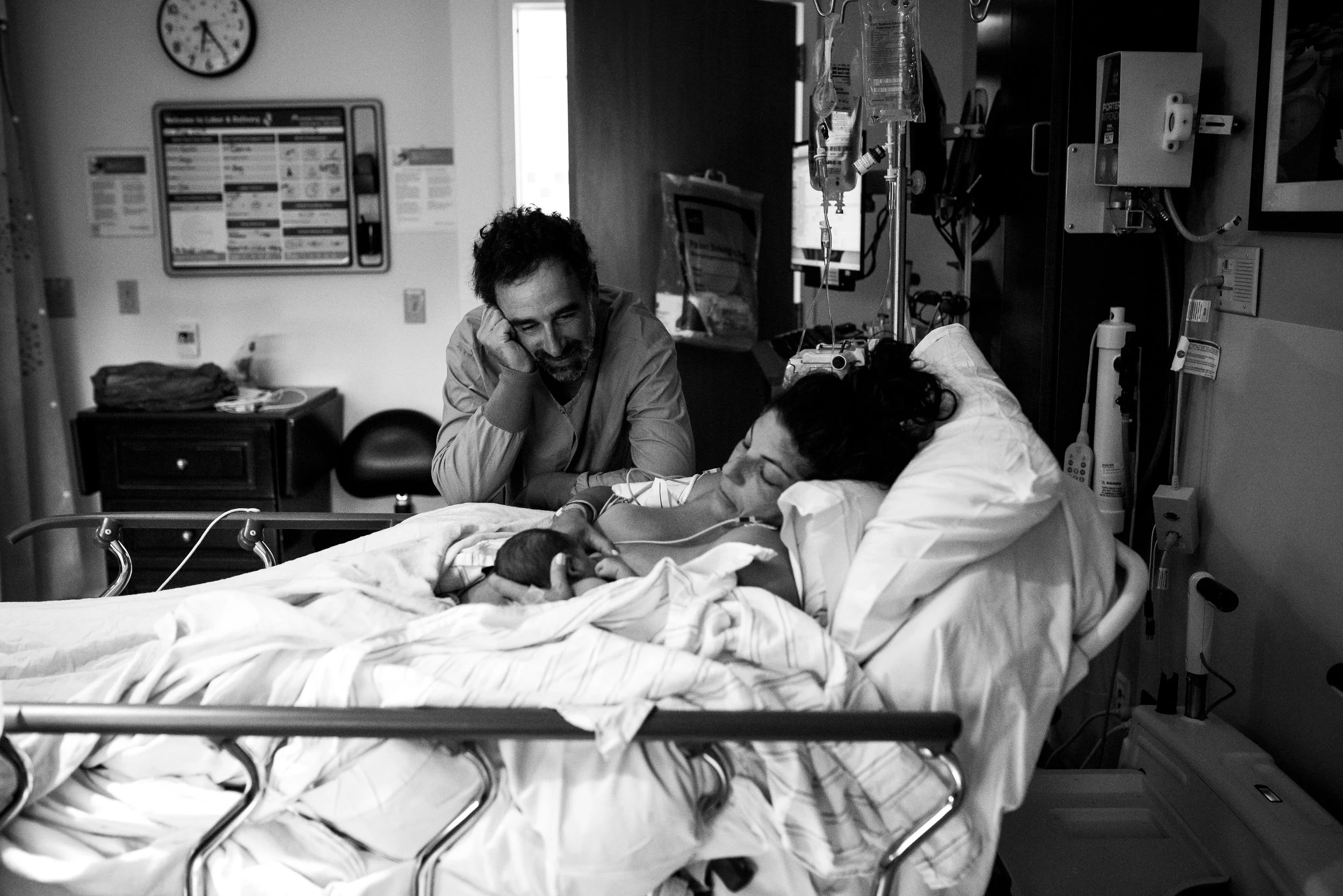 In a hospital room, a woman is lying in a bed connected to medical equipment, with a man sitting beside her, smiling and talking to her. The room has medical monitors, IV drip, and equipment in the background.
