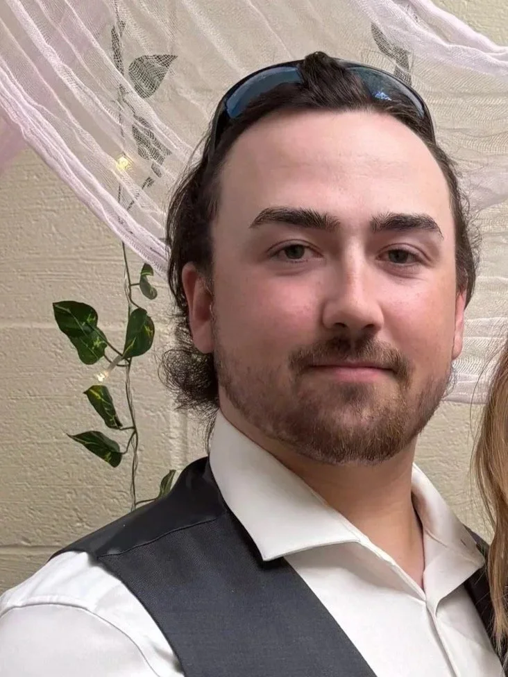 A young man with a beard and mustache smiling, wearing a beige t-shirt, a silver chain necklace, and a baseball cap, standing outdoors on a paved walkway with green grass, benches, and trees in the background.