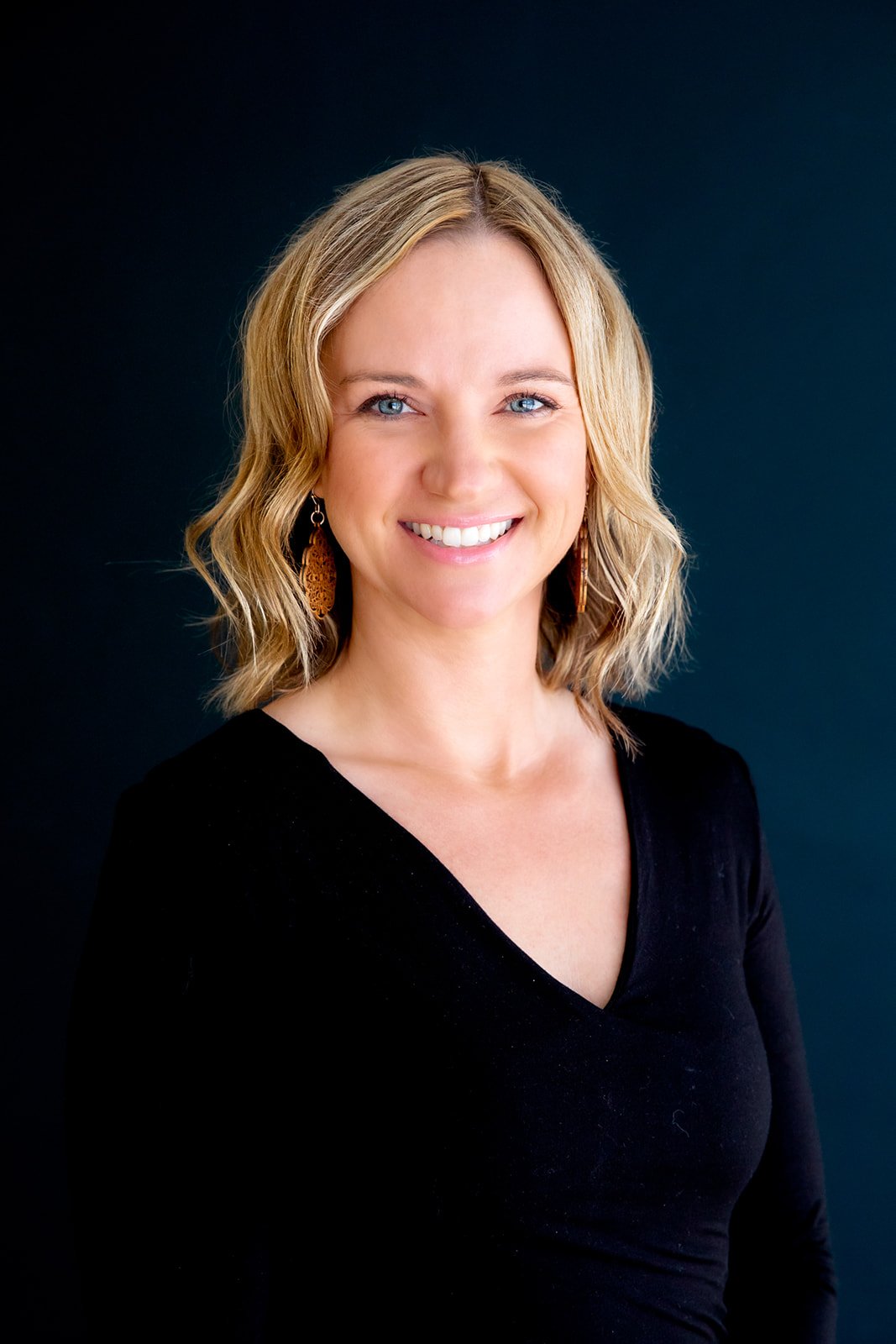 Woman smiling in a black top with medium-length blonde hair against a dark background.