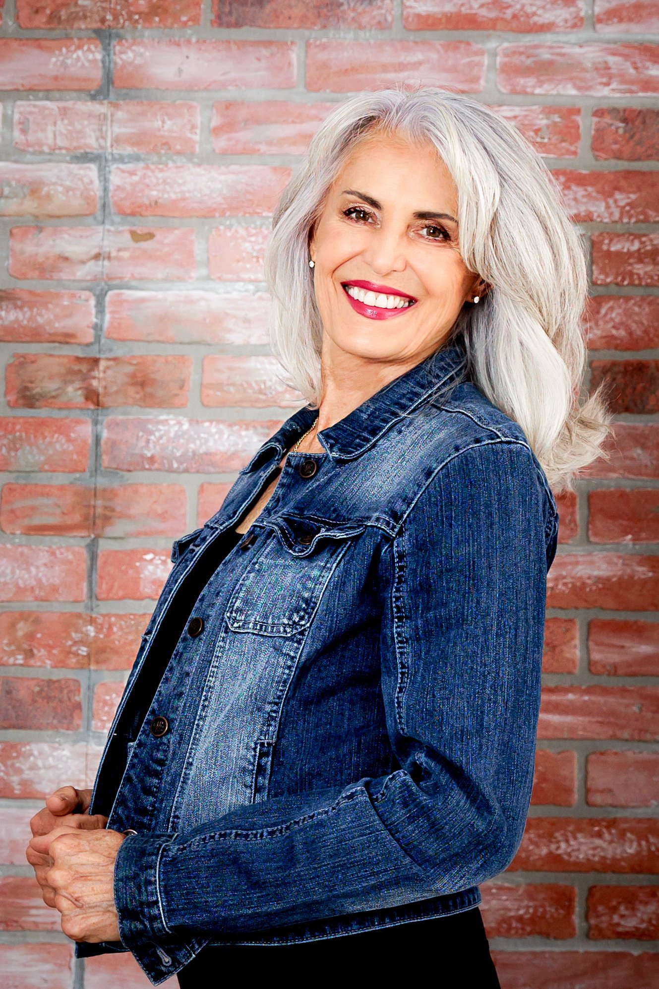 Smiling woman with gray hair wearing a denim jacket against a brick wall.