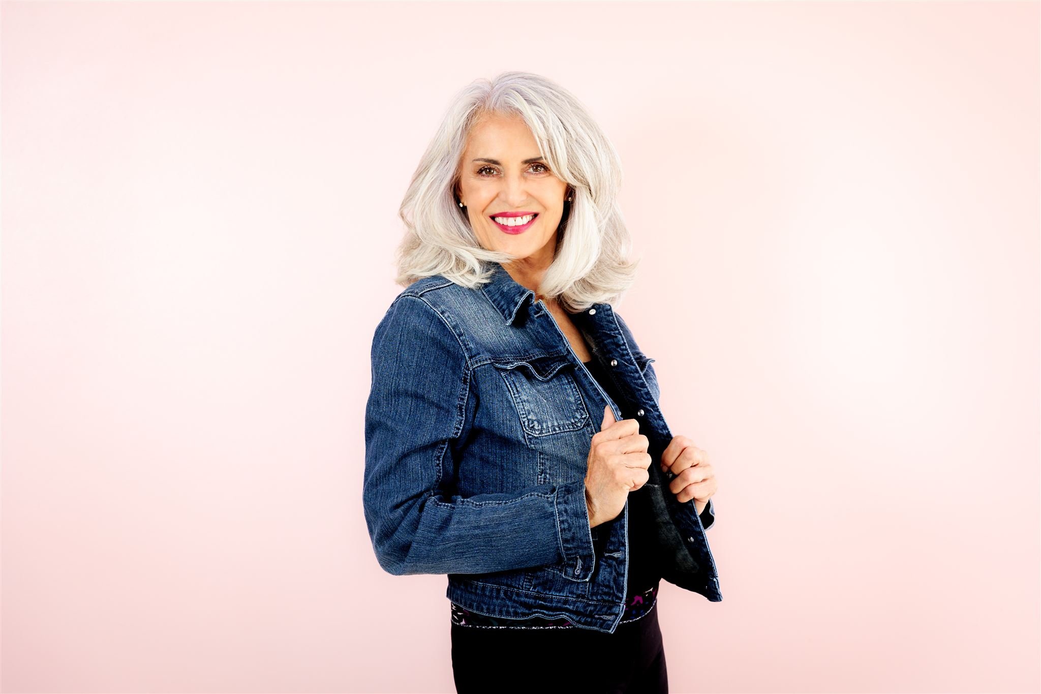 Smiling woman with gray hair wearing a denim jacket against a light pink background.