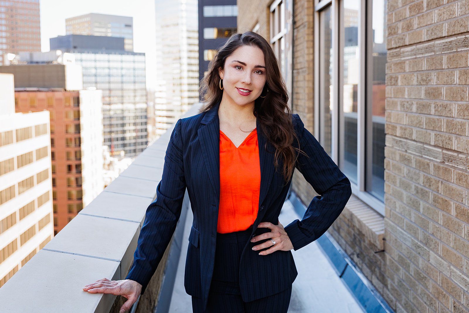 Woman in navy suit with orange blouse standing on rooftop with city buildings in the background.