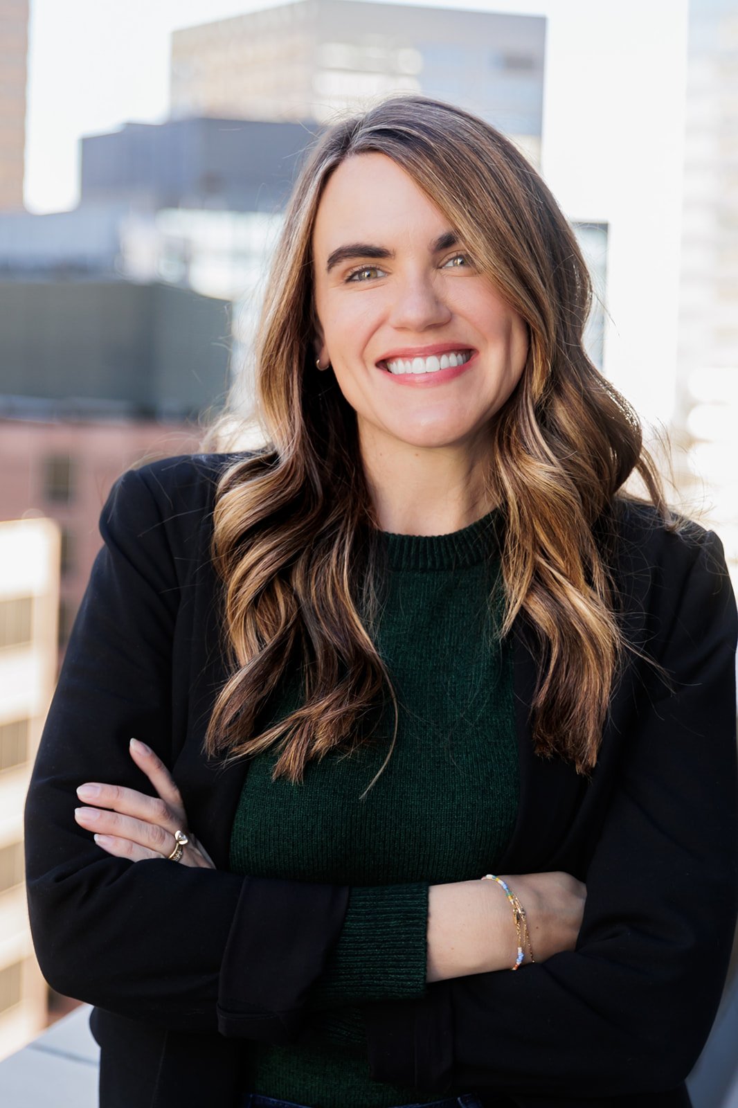 Smiling woman with long hair and wearing a dark green sweater and black blazer, standing with arms crossed in an urban setting.
