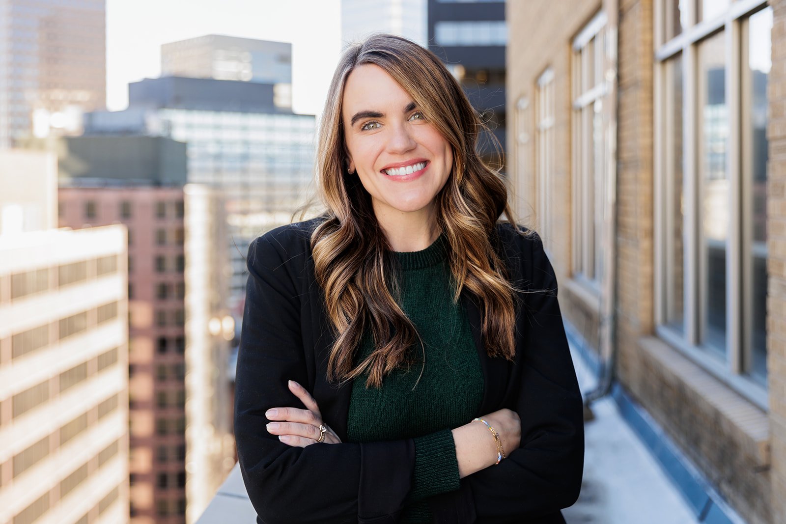Smiling person with long hair standing on a rooftop terrace with city buildings in the background.