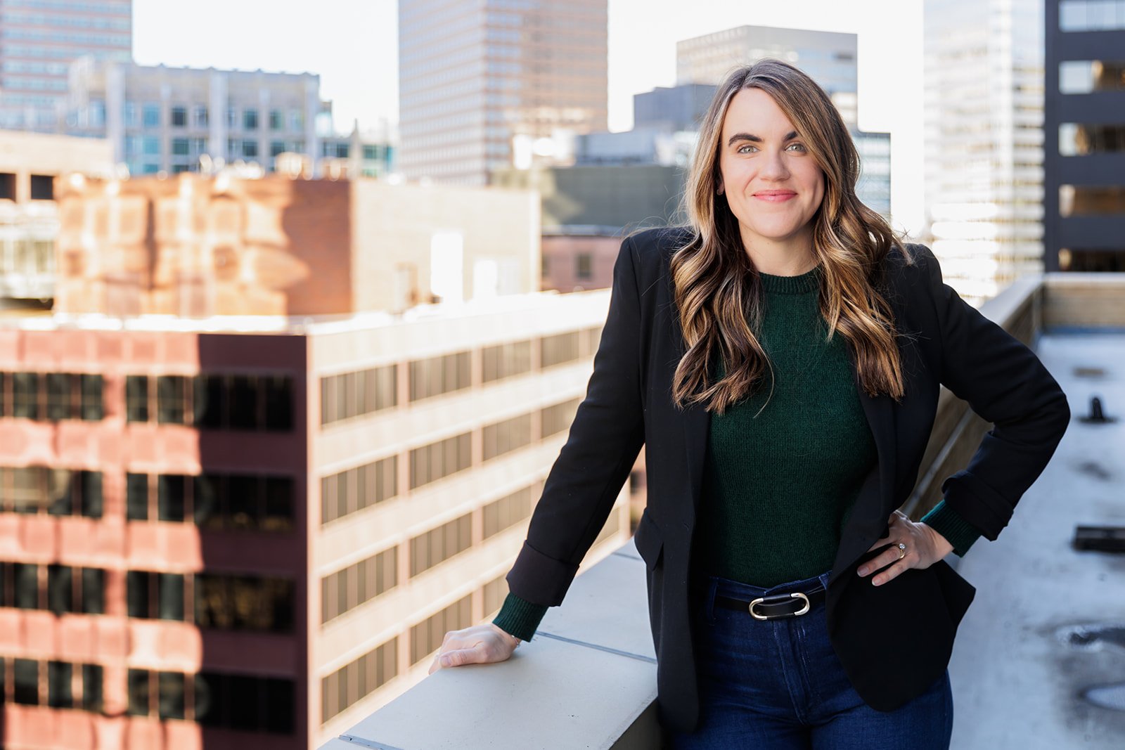 Woman standing on a rooftop with city buildings in the background, wearing a black blazer and green sweater.