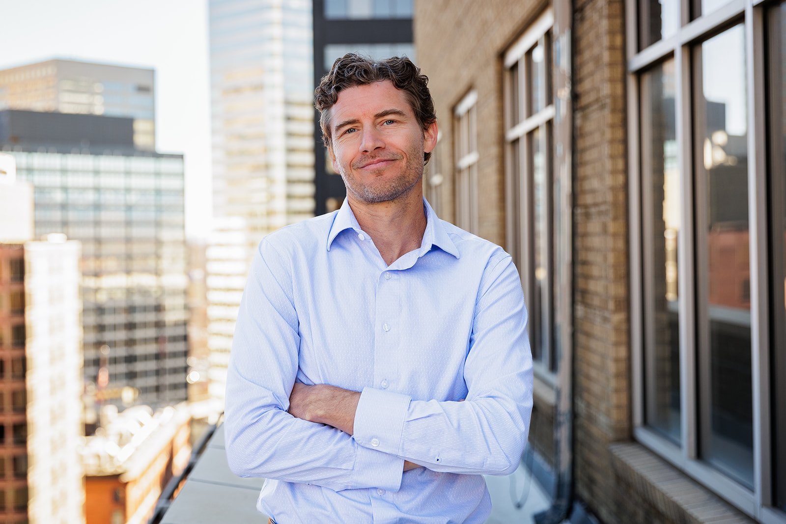 Man in a light blue shirt standing with arms crossed on a city rooftop with skyscrapers in the background.