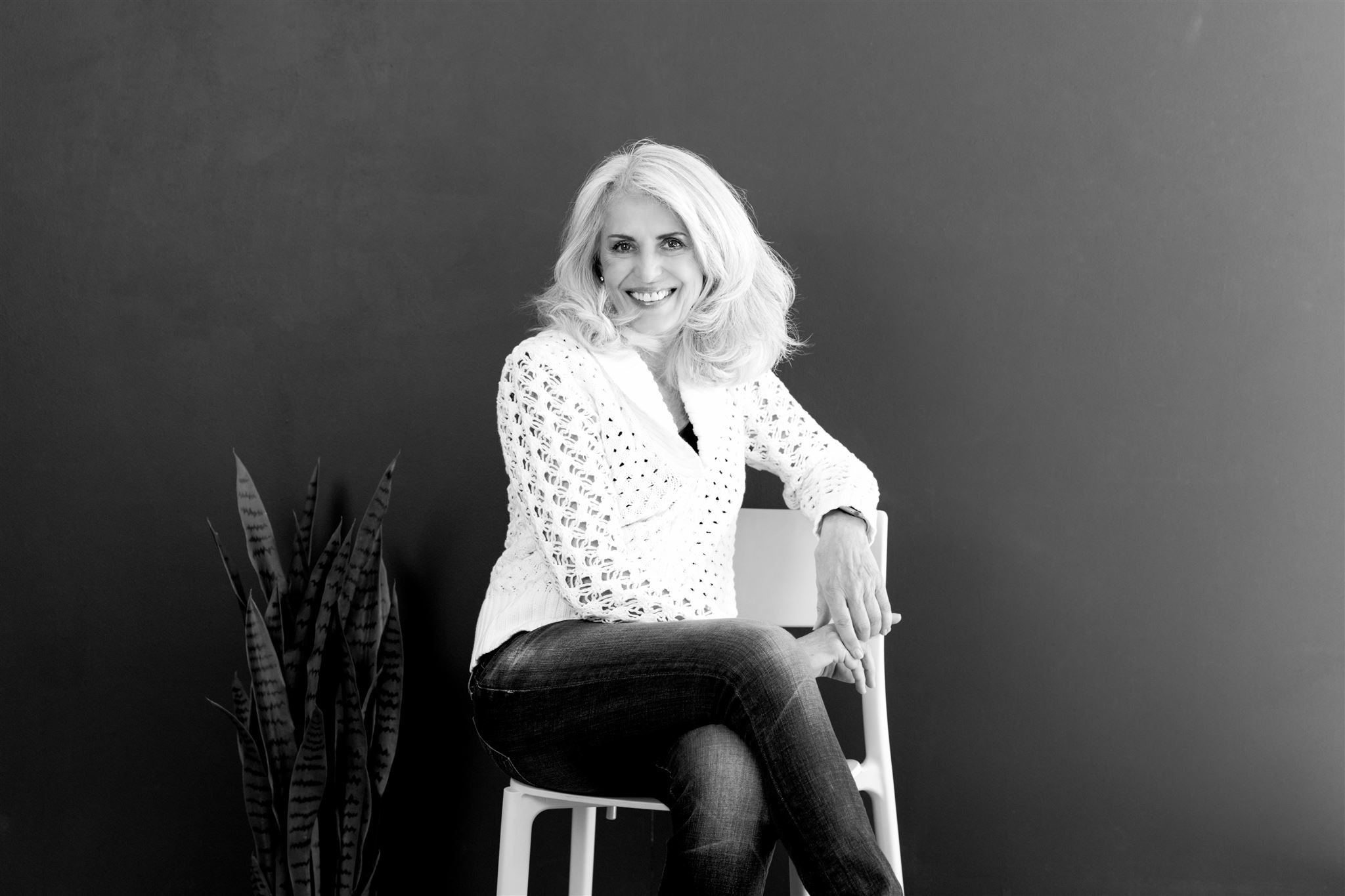 Black and white photo of a woman with shoulder-length hair sitting on a chair, wearing a knit sweater and jeans, next to a tall plant.