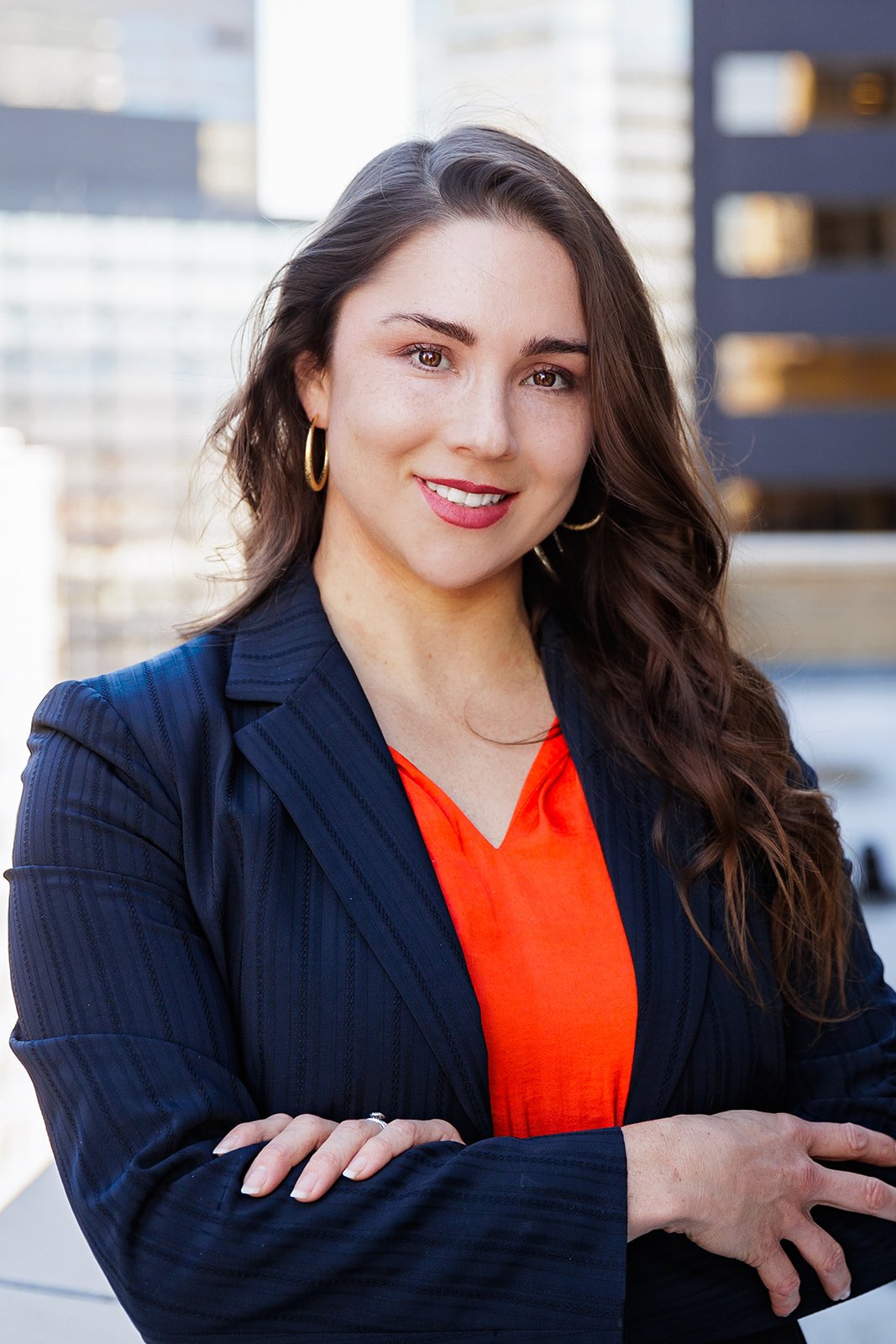 Smiling woman in a professional outfit posing with arms crossed, urban background.