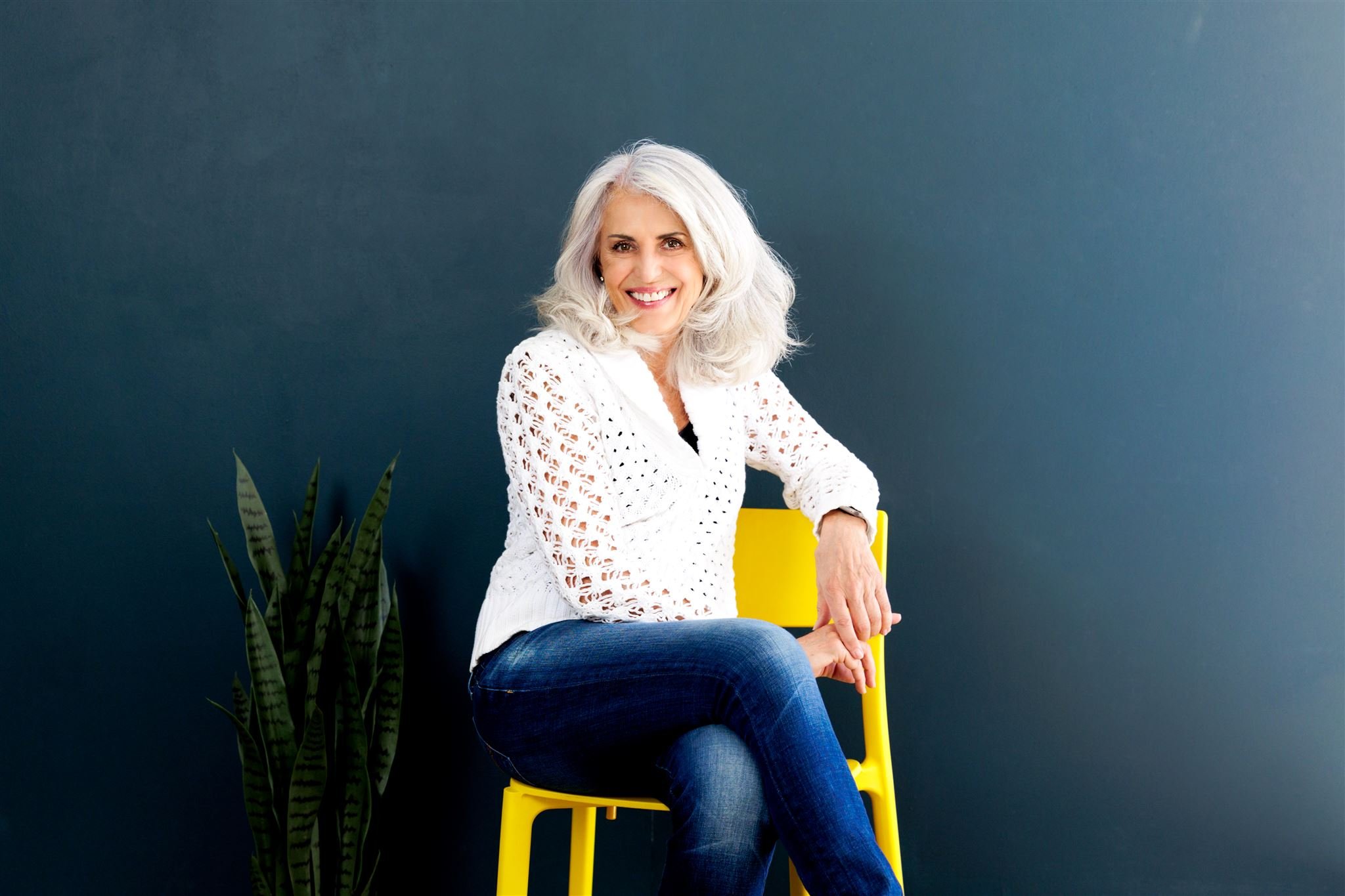 A woman with long gray hair in a white knit sweater sits on a yellow chair in front of a dark teal background, next to a potted snake plant.