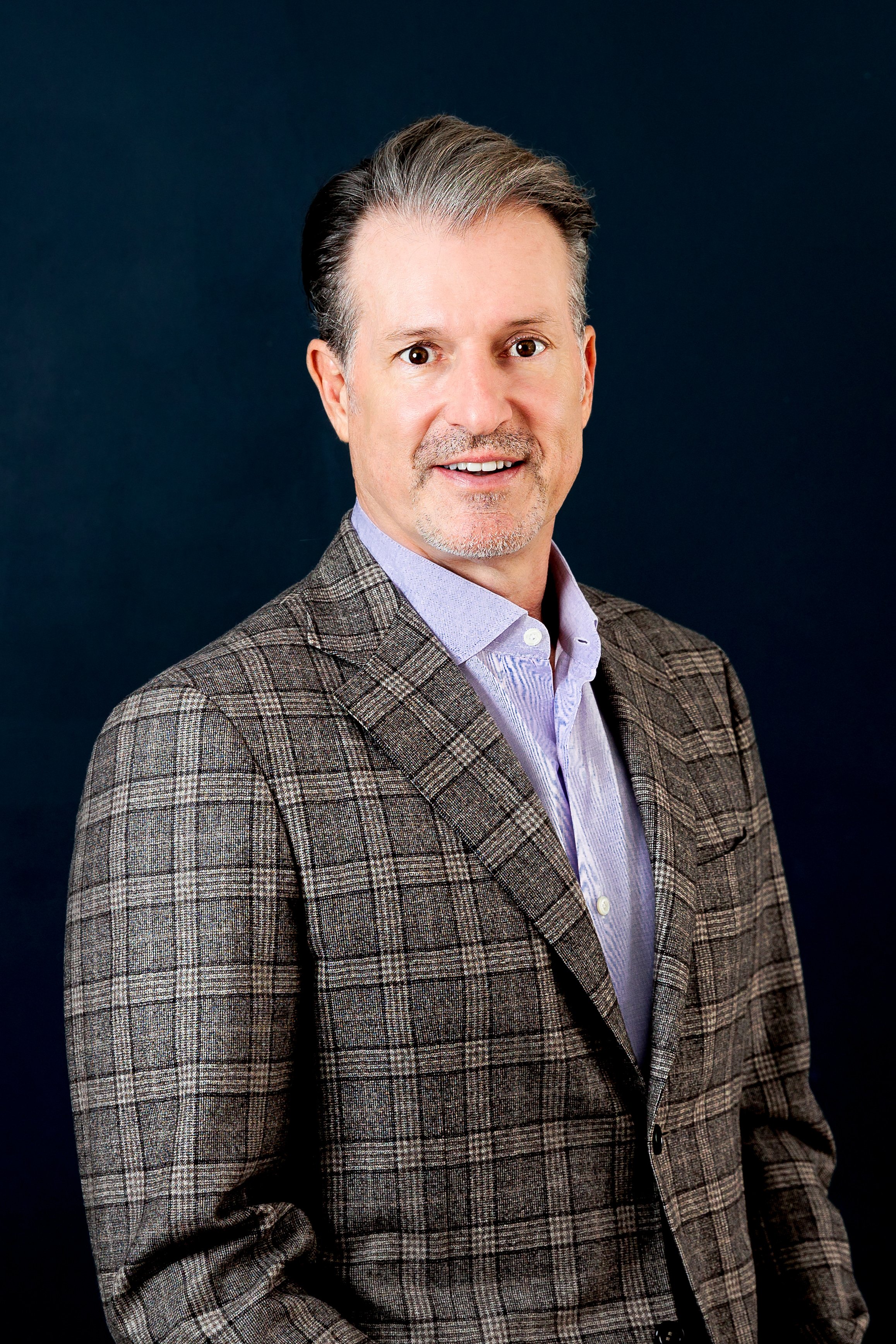 Man in a plaid blazer and light shirt posing against a dark background.