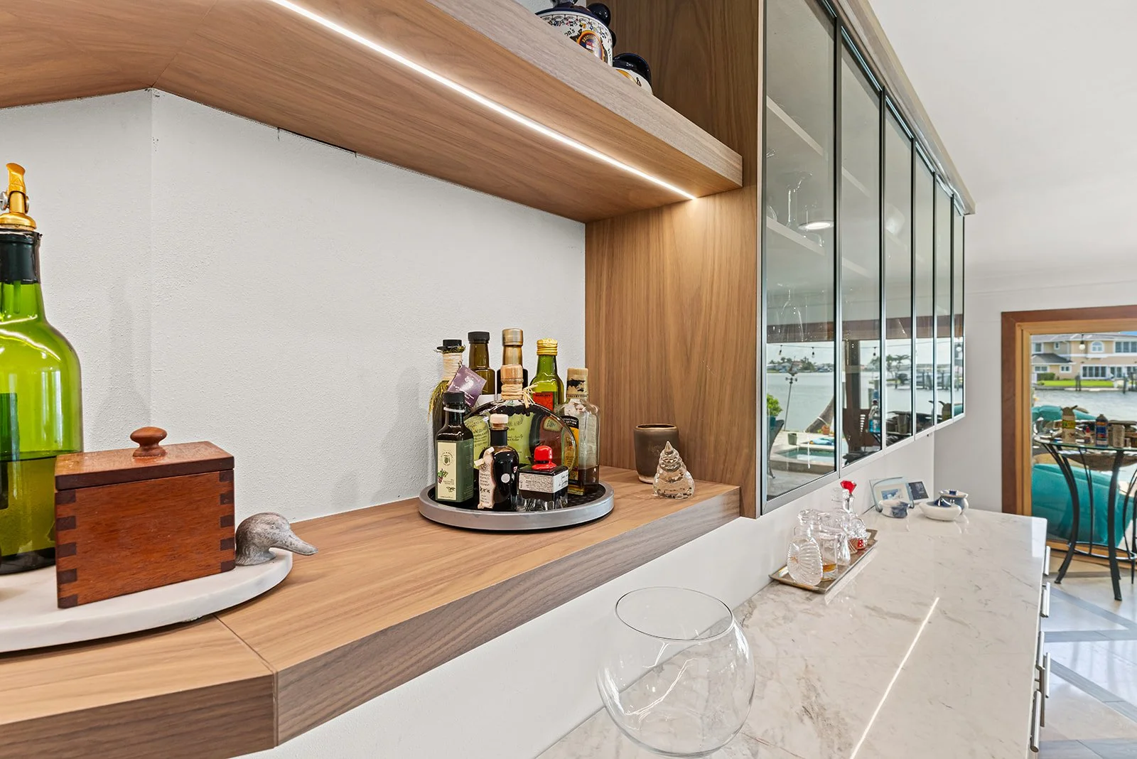 A kitchen counter with various bottles of alcohol and seasonings on a round tray. There are decorative items, including a small sculpture and a wooden box, on a white marble surface. A large window shows an outdoor scene with boats on water.