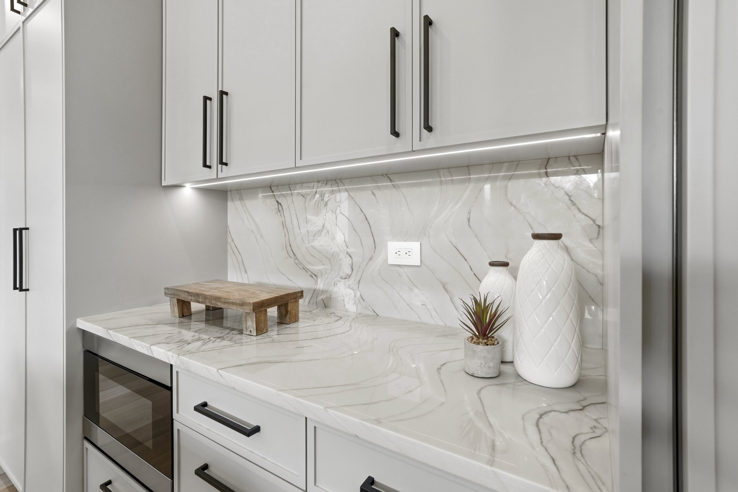 Modern kitchen with white cabinets, black handles, marble countertops and backsplash, a small wooden cutting board, and decorative white vases with plants.