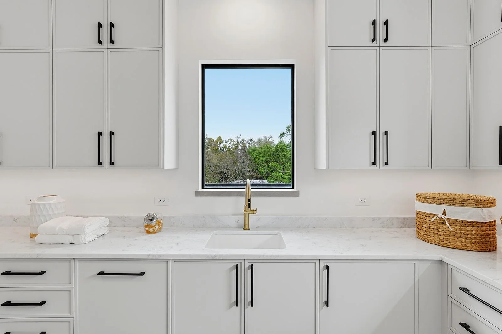 White kitchen with white cabinets, a marble countertop, and a window overlooking trees. There is a gold faucet, folded towels, a decorative vase, and a woven basket on the counter.