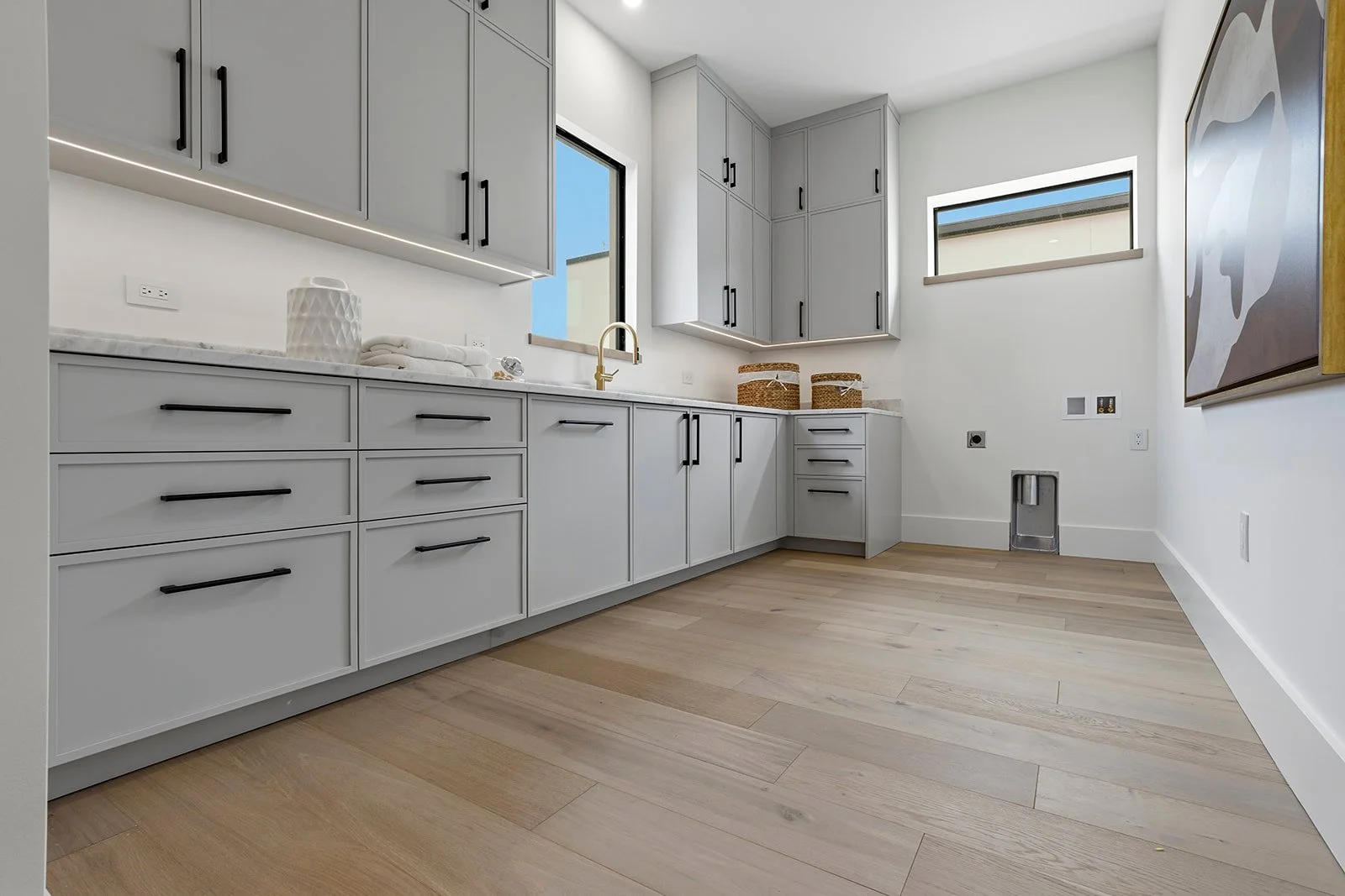 Empty laundry or utility room with white cabinets, light wood flooring, and a window, with built-in space for appliances and a gold faucet at the sink.