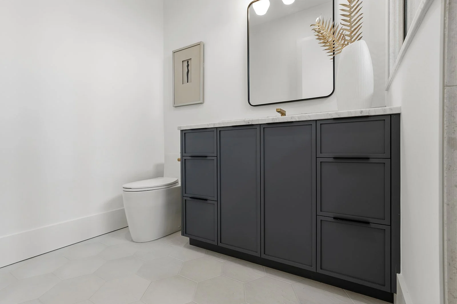 Modern bathroom with a white wall and light gray hexagonal tile floor. Features a dark gray vanity with a white marble countertop, a rectangular mirror, and decorative items including a vase with dried leaves. A white toilet is positioned next to the