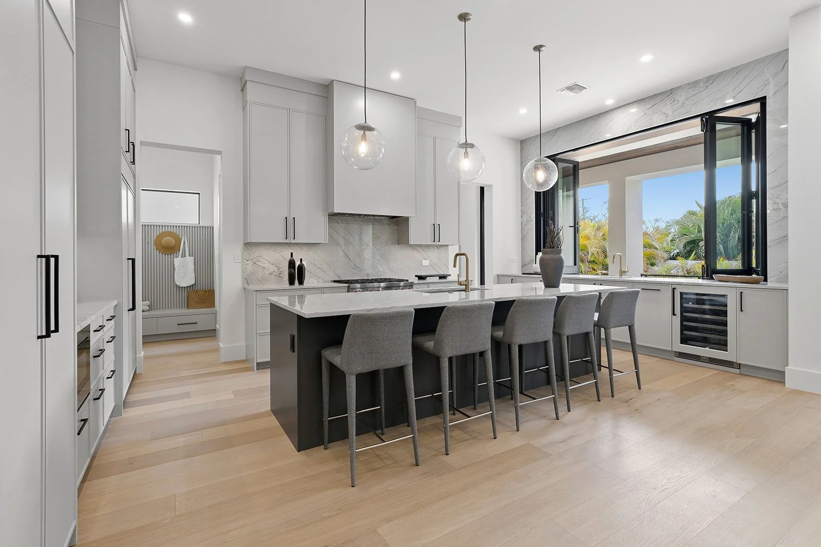 Modern kitchen with white cabinets, a black island with gray bar stools, marble backsplash, large windows, and light wooden flooring.