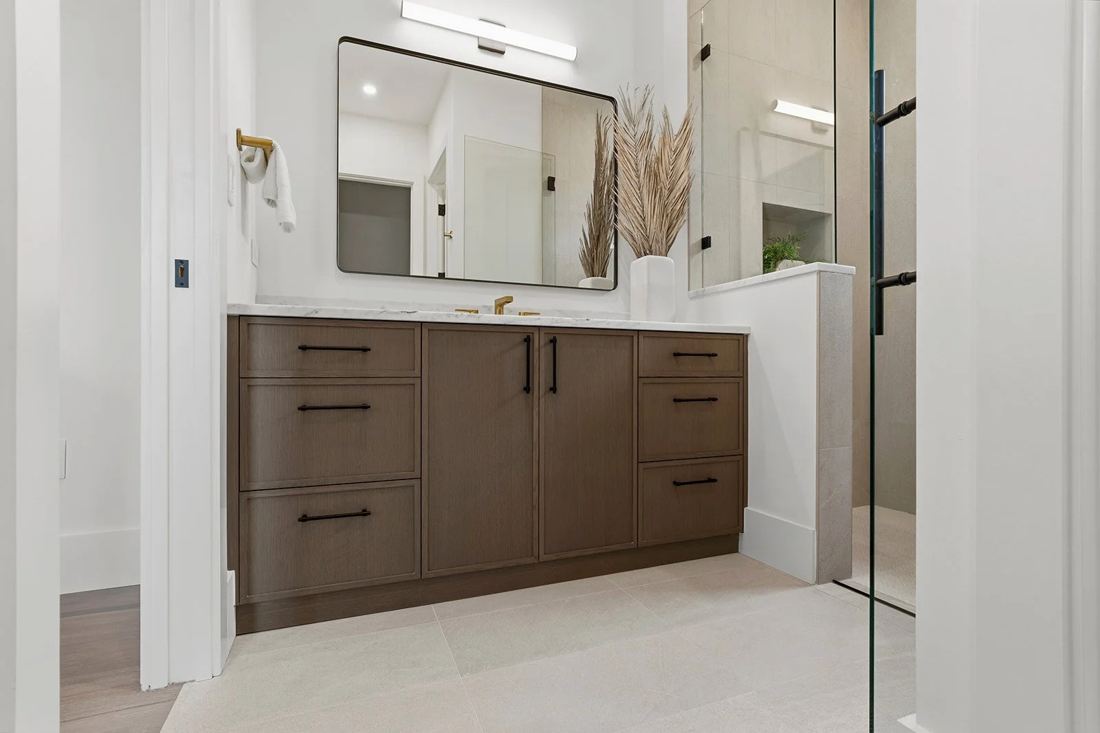 Modern bathroom vanity with brown cabinets, a white marble countertop, gold faucet, large mirror, decorative dried plants, and a walk-in shower with glass door.