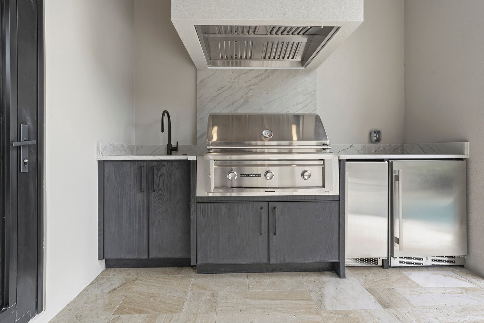 Modern outdoor kitchen with stainless steel grill, black cabinets, white marble backsplash, small sink, and mini refrigerator.